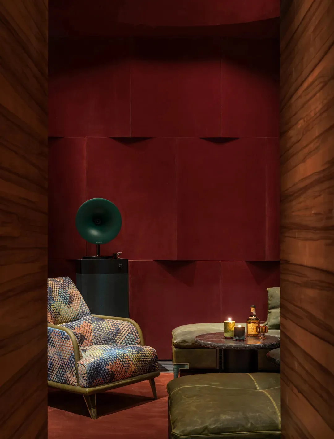 Intimate lounge corner with deep red textured walls, walnut wood paneling, a multicolored patterned armchair, green gramophone horn, and leather ottoman with drinks table.
