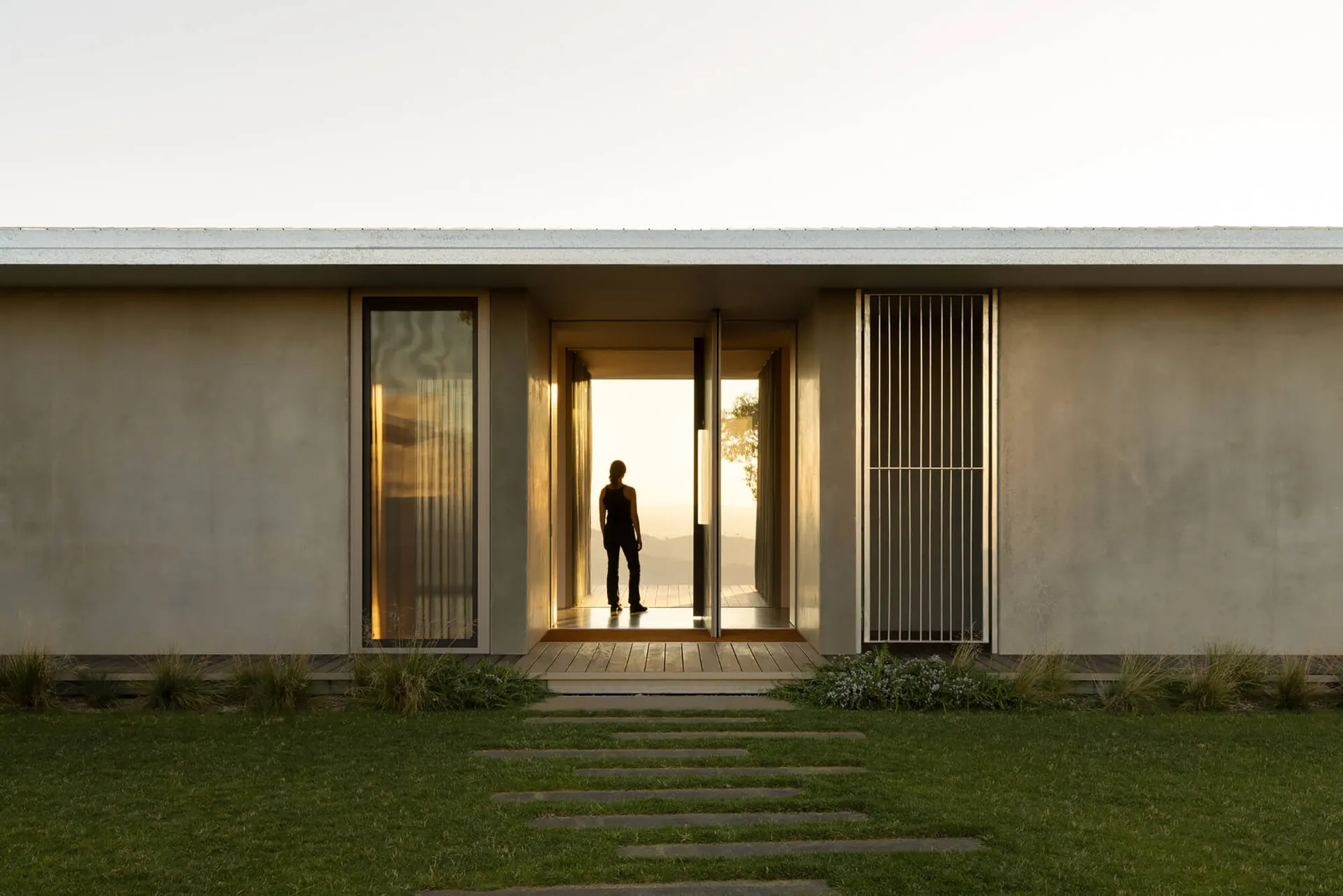 Modern minimalist concrete house with large glass doors and vertical metal screen, golden sunset light illuminating an open courtyard entrance with a woman standing on the wooden deck overlooking distant hills.