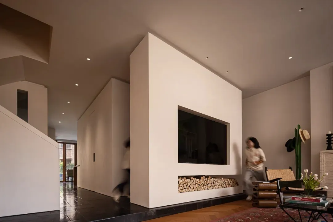Modern minimalist living room featuring a built-in TV and fireplace wall with stacked firewood, oak herringbone flooring, and neutral beige walls.