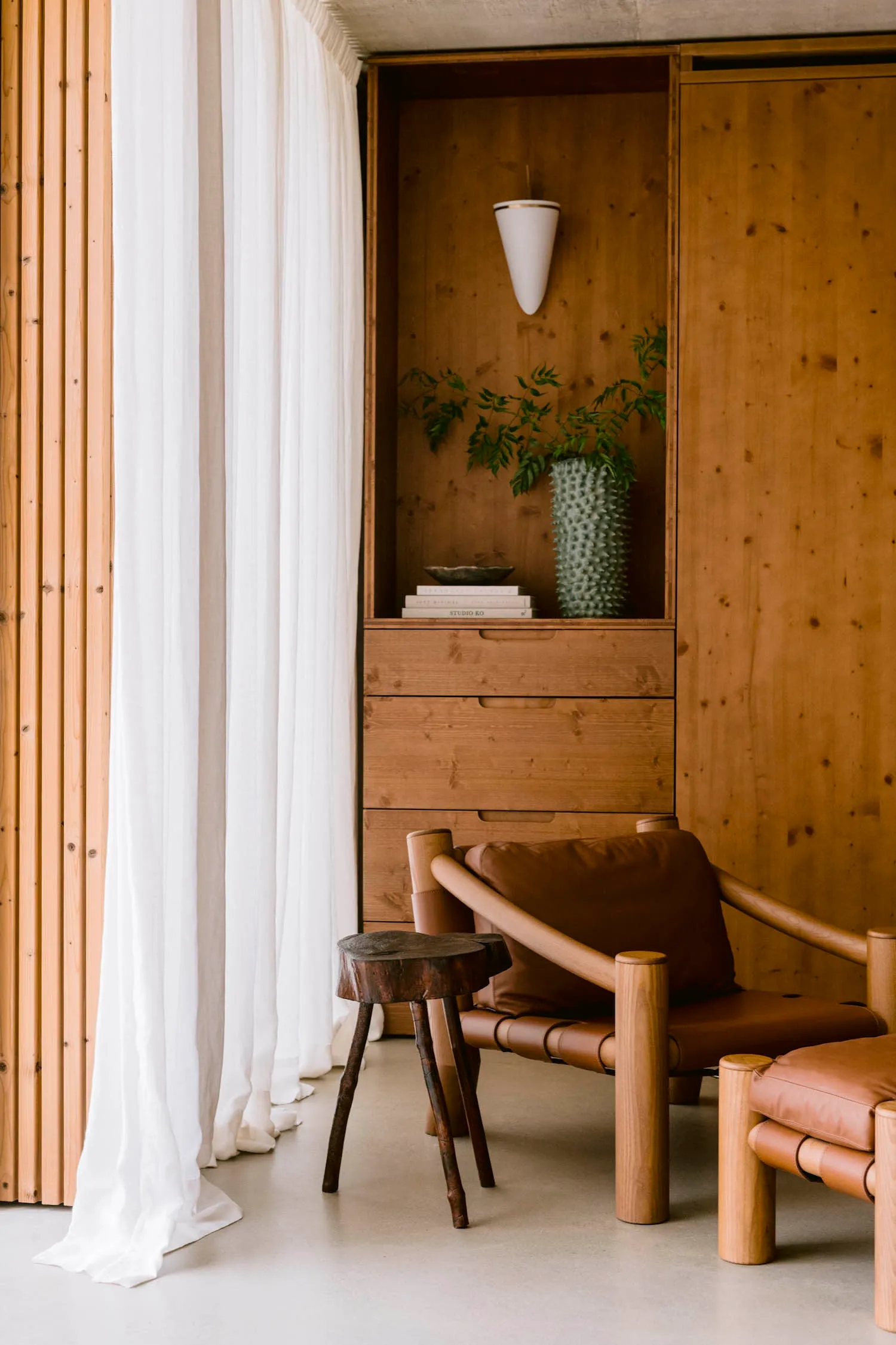 Modern minimalist living room corner featuring a brown leather and oak armchair, dark wood stump side table, built-in oak shelving with books and plants, white sheer curtain, and concrete ceiling.