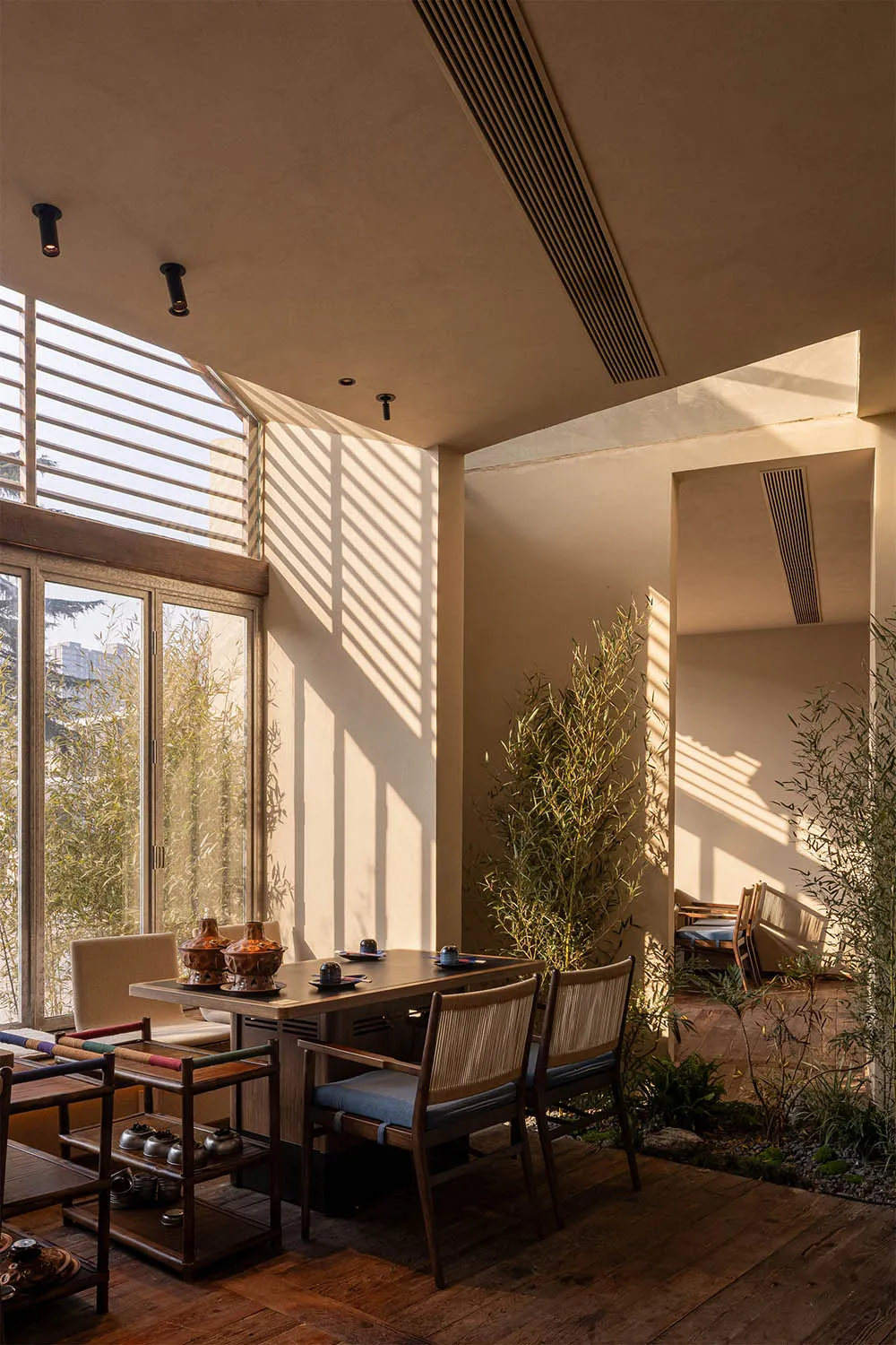 A tranquil dining area featuring a dark wood table, woven rattan and fabric dining chairs, colorful wooden side tables, and tall bamboo plants, with strong sunlight casting striped shadows on beige plaster walls.