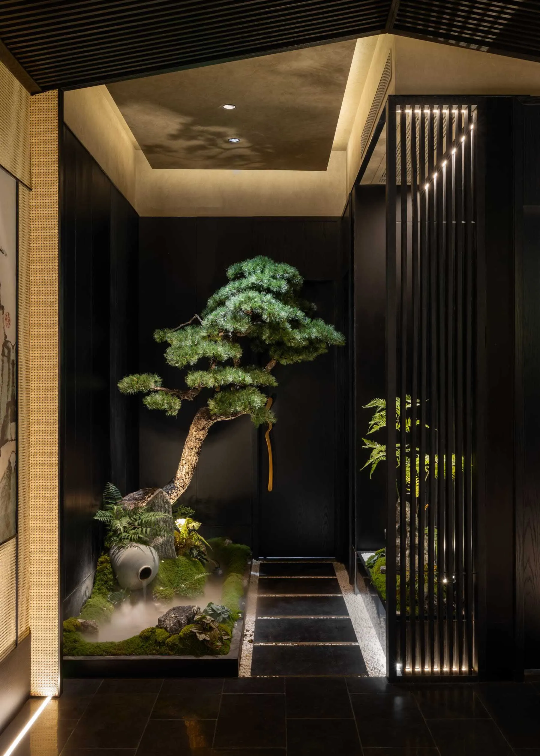 Modern indoor Japanese garden entrance with large bonsai pine tree, moss garden, stone water feature emitting mist, dark wood paneling, and vertical illuminated wooden slats.
