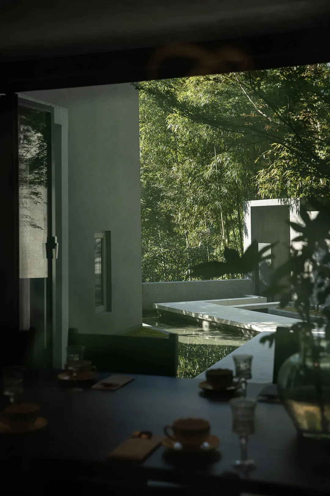 Modern minimalist dining area with dark table set, looking out through large openings to a concrete courtyard with water pond and dense bamboo forest.