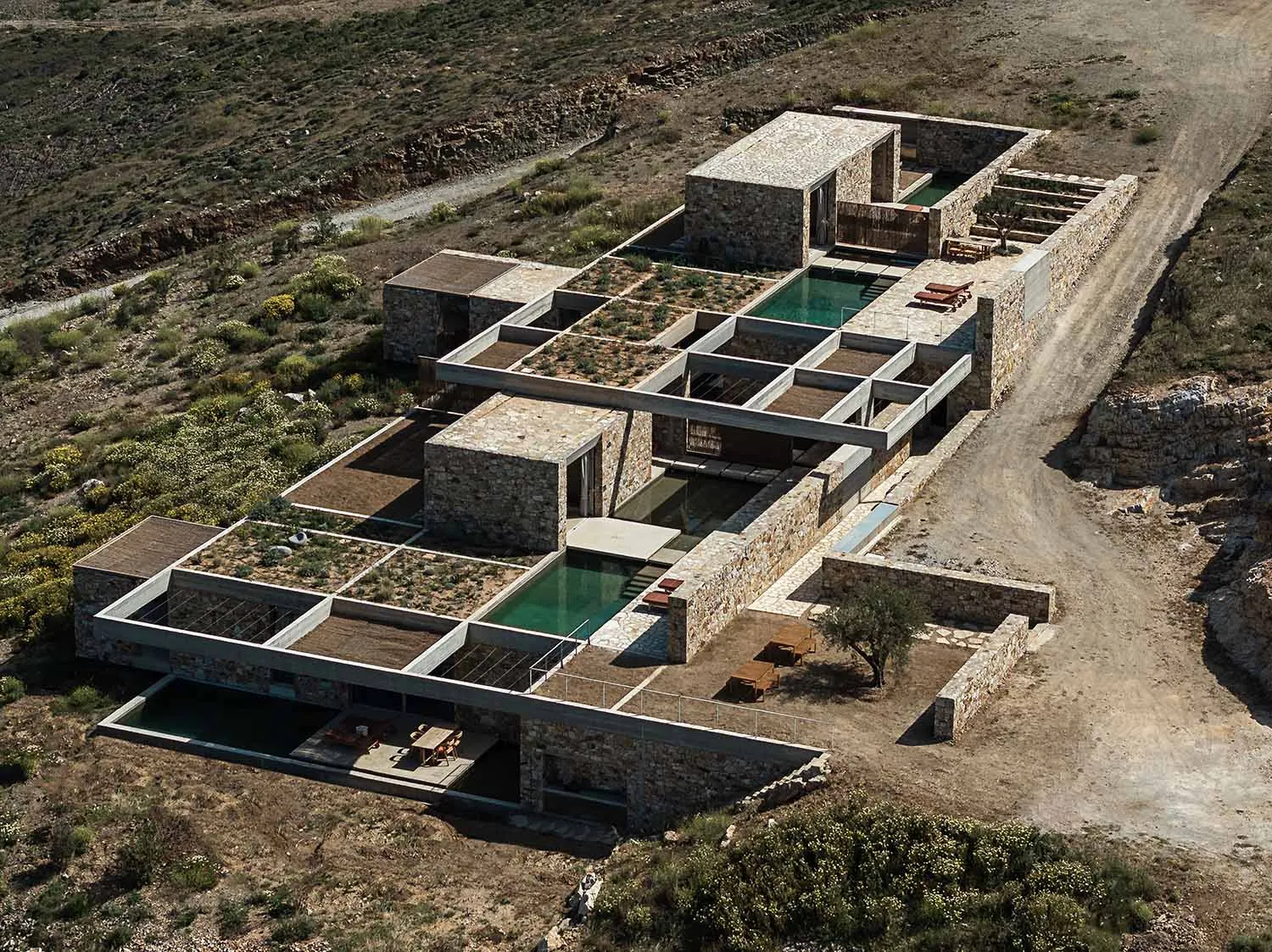Aerial photograph of a modern stone courtyard villa with green infinity pools integrated into the arid Mediterranean hillside.