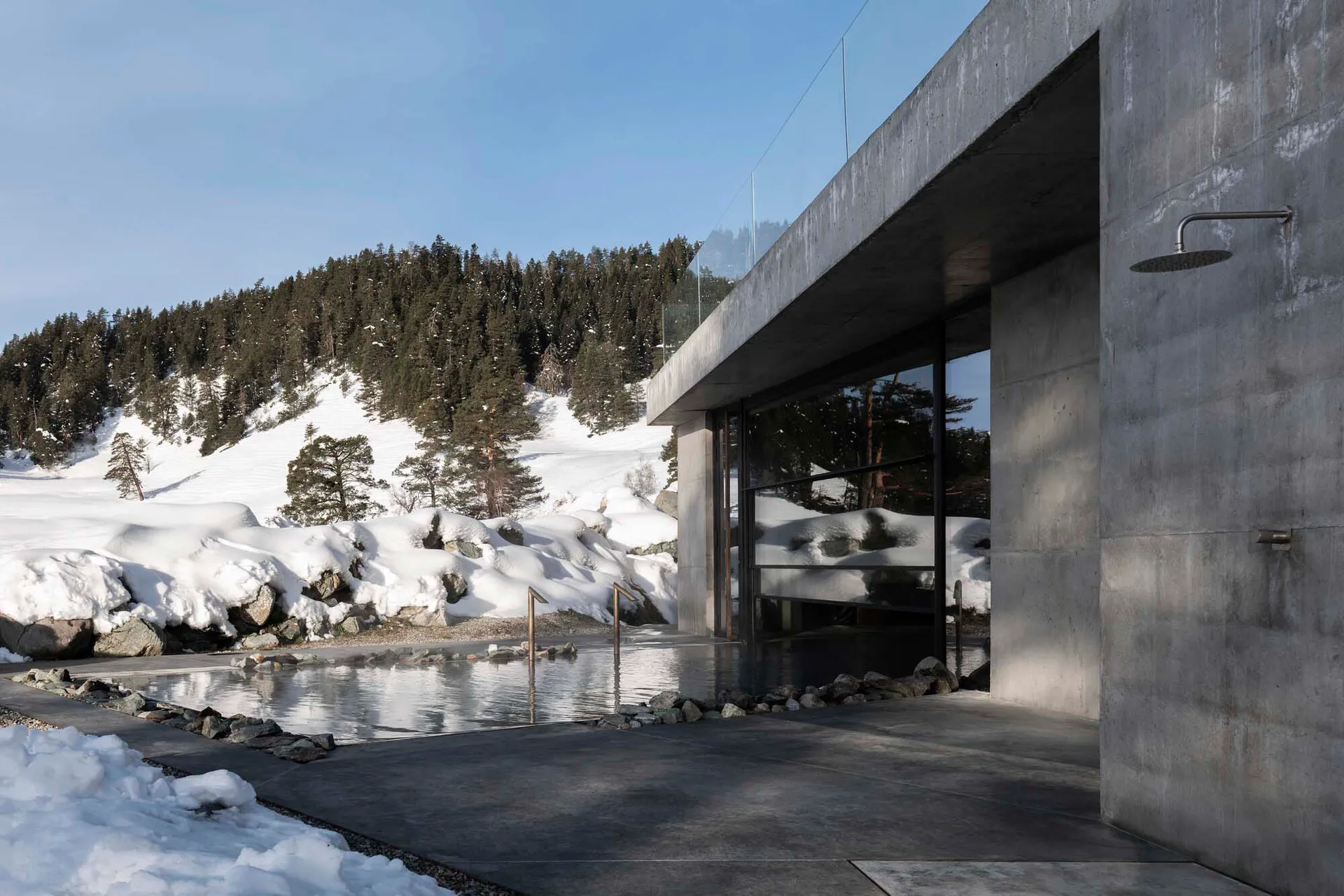Modern concrete outdoor hot spring bath with glass walls, set beside a snowy mountain and forest in winter sunlight.