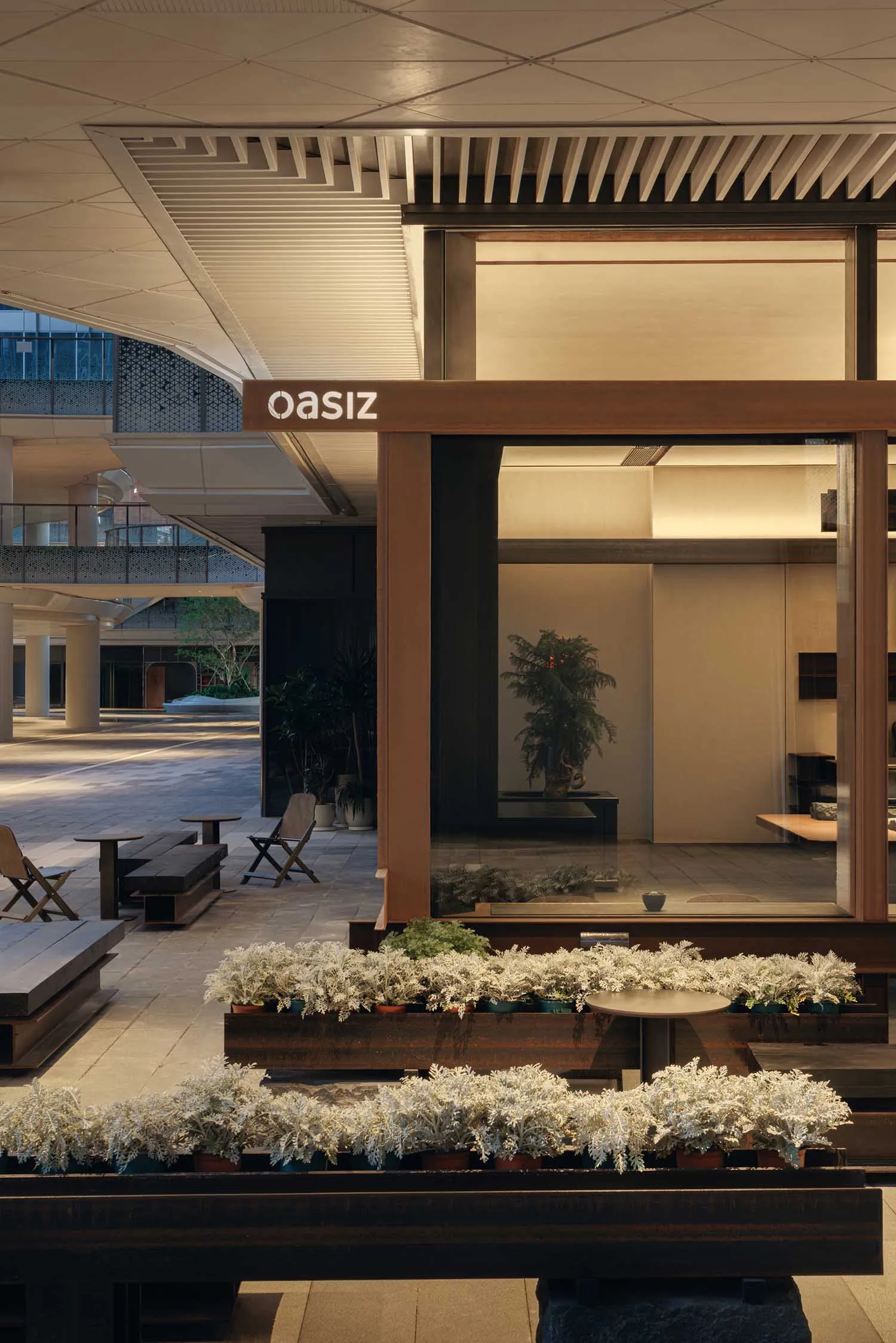 Modern courtyard lounge with corten steel planters filled with silvery white foliage, wooden benches, and a large glass-walled room in the background under a covered architectural ceiling.