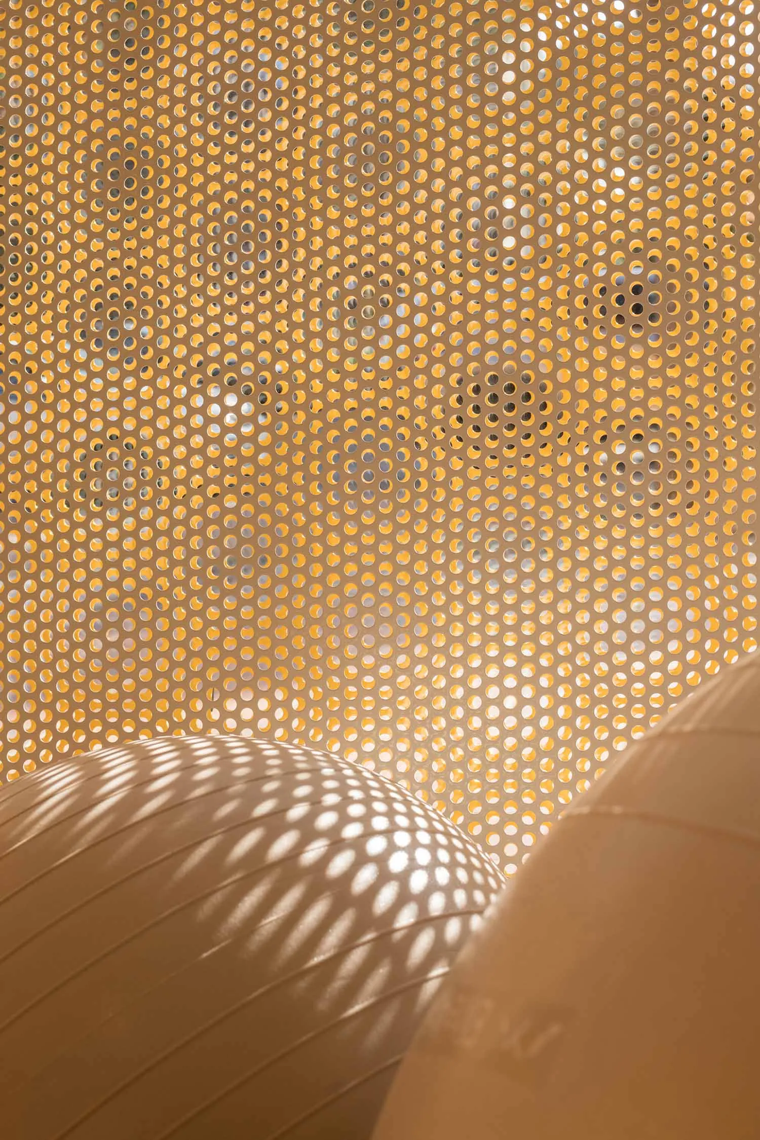 Close-up view of a light-colored wooden perforated screen with hundreds of uniform circular holes, with bright sunlight creating dotted shadow patterns on a curved wooden surface below.