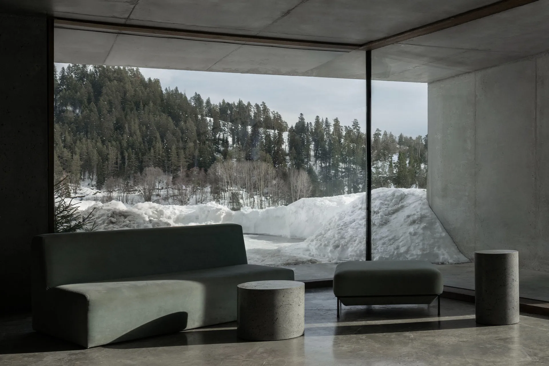 Modern minimalist living room with concrete walls and floor, featuring a curved green fabric sofa, matching ottoman, cylindrical concrete side tables, and a massive picture window overlooking a snowy forest and mountain.