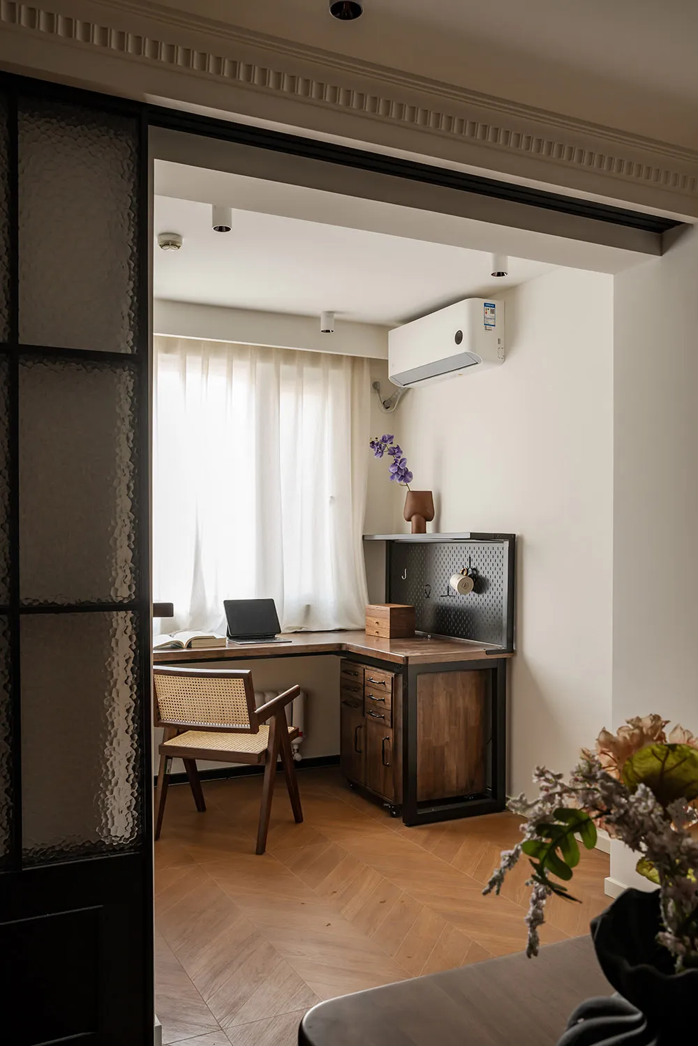 View into a compact home office with an L-shaped walnut desk, cane chair, pegboard organizer, and large window with sheer curtains.