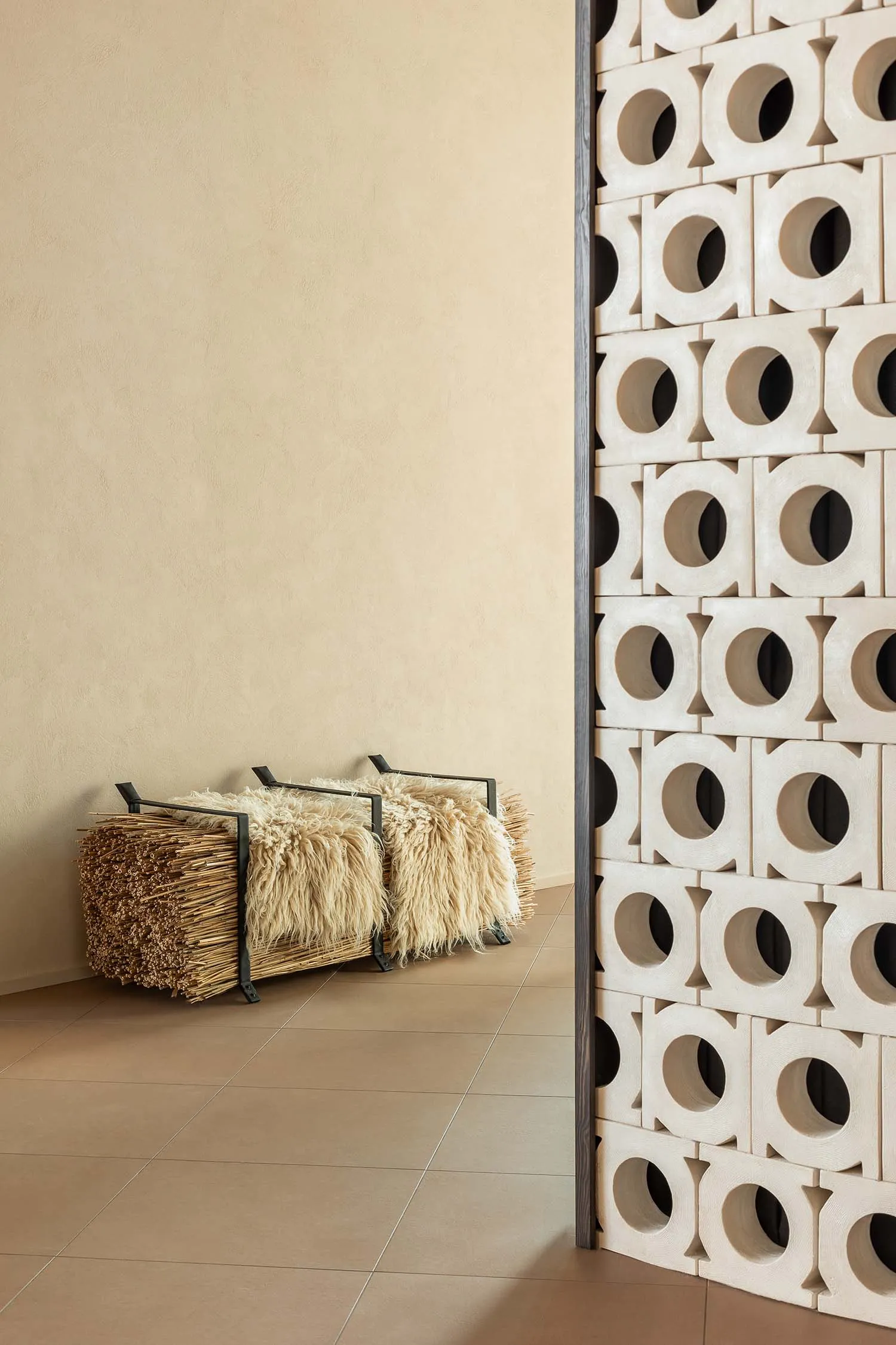 Modern minimalist entryway featuring a large off-white perforated terracotta screen, a black metal-framed bench with two cream sheepskin throws, and a bundle of dried reeds against a textured beige wall and large-format beige floor tiles.