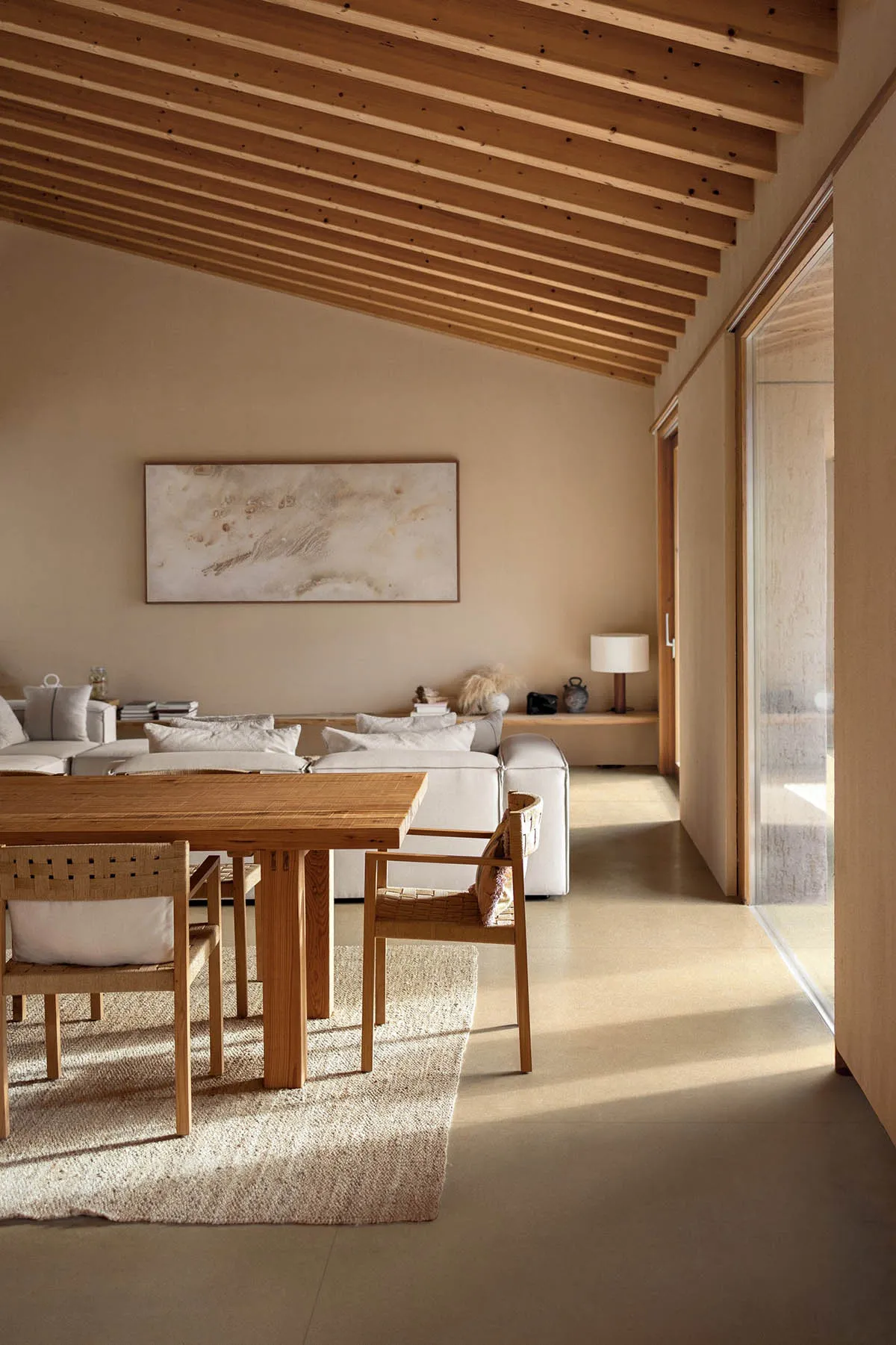 Modern minimalist dining area with solid oak table and woven rattan chairs on a textured rug, white sectional sofa in the background, exposed wooden beam ceiling and large abstract painting on the wall.
