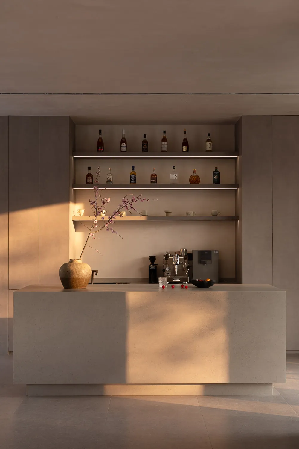 Modern minimalist concrete kitchen island used as a home bar with backlit floating shelves displaying liquor bottles and tableware, featuring a large ceramic vase with flowering branches in soft natural light.