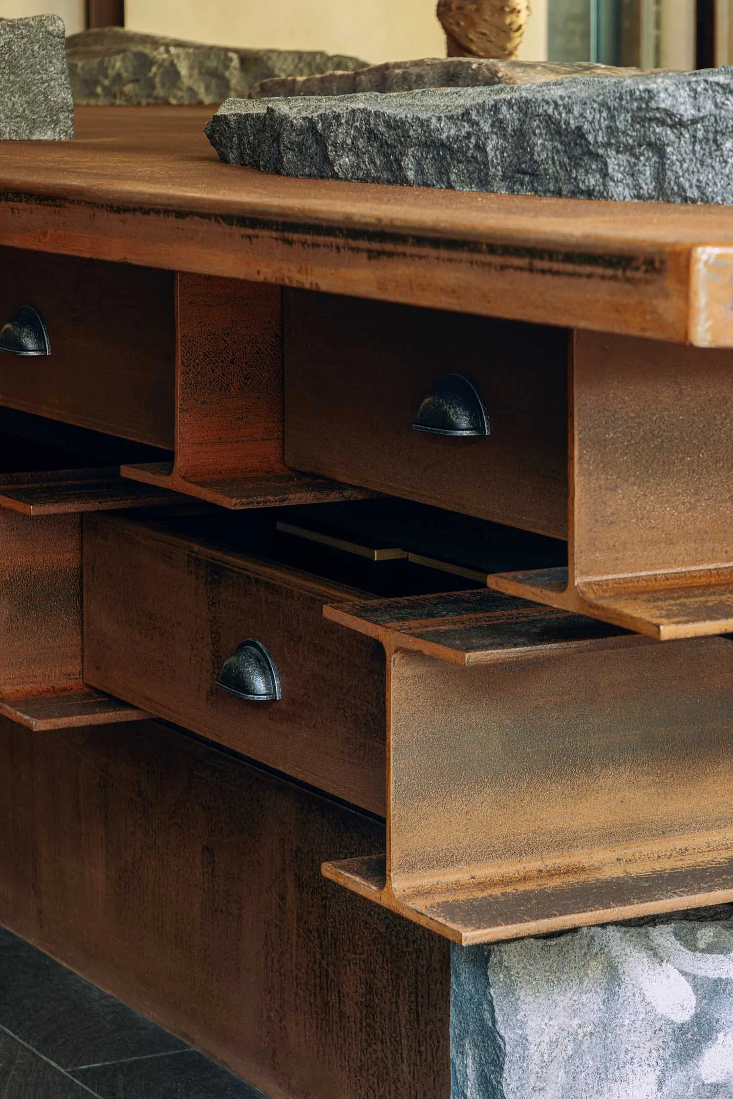 Close-up of a custom bathroom vanity featuring a thick weathered steel frame with integrated drawers, topped by a massive rough-hewn stone basin, resting on a natural stone base.