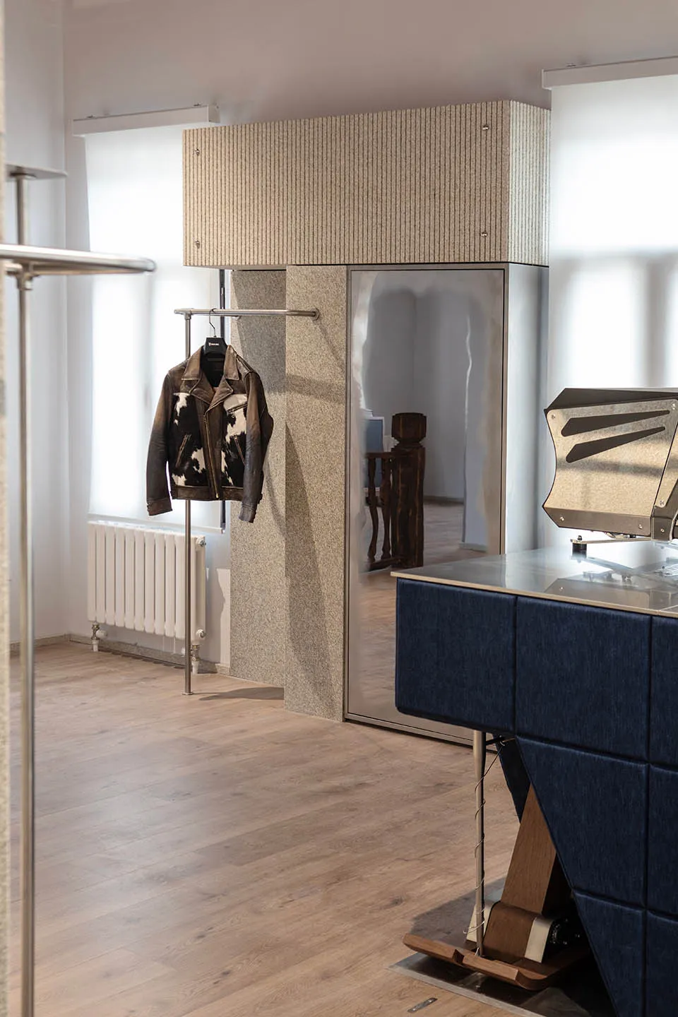 Modern minimalist entryway featuring a tall fluted stone wardrobe with integrated mirror, a blue upholstered console table, and a leather jacket hanging on a chrome rack against a light wood floor.