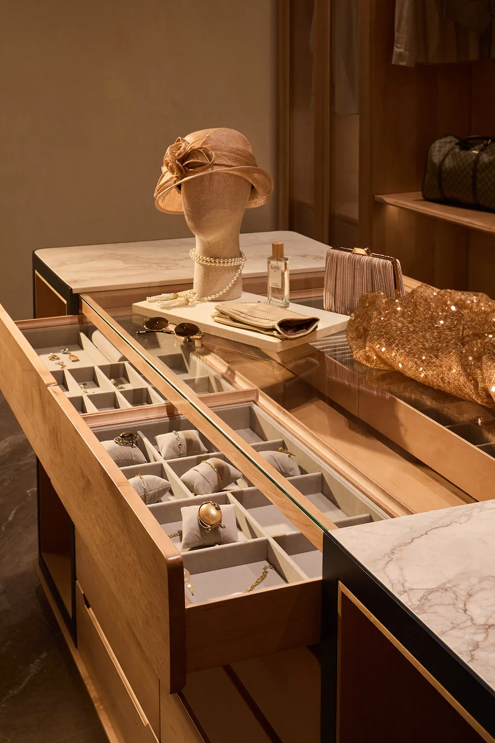 Luxury dressing room featuring a custom oak and marble jewelry display island with multiple open drawers revealing organized fine jewelry, pearl necklace on a mannequin wearing a beige hat, perfume bottle, and gold accessories under warm lighting.