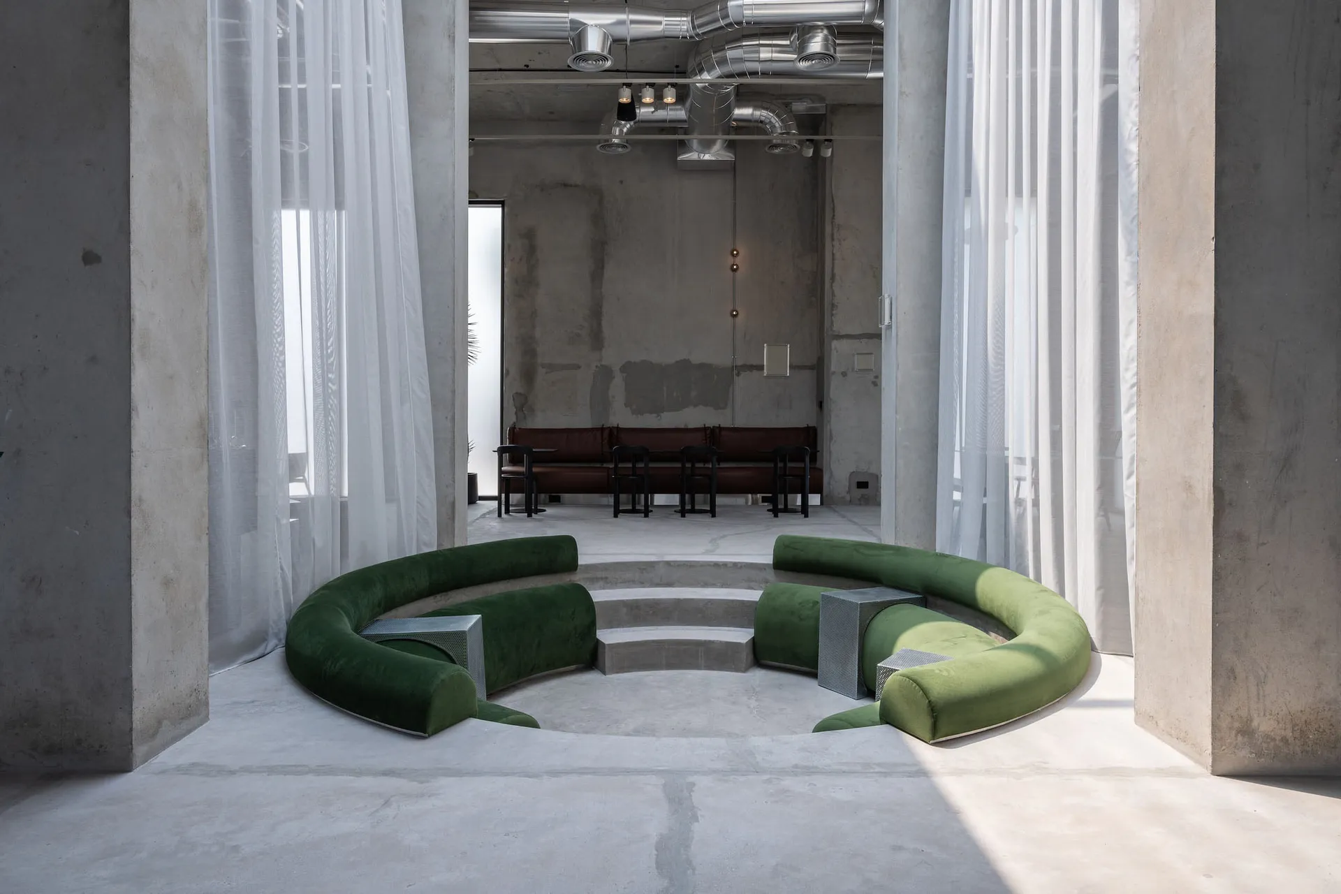 Modern brutalist interior featuring a large curved emerald green velvet sofa arranged in a sunken circular platform, surrounded by sheer white curtains and exposed concrete walls with visible ductwork.