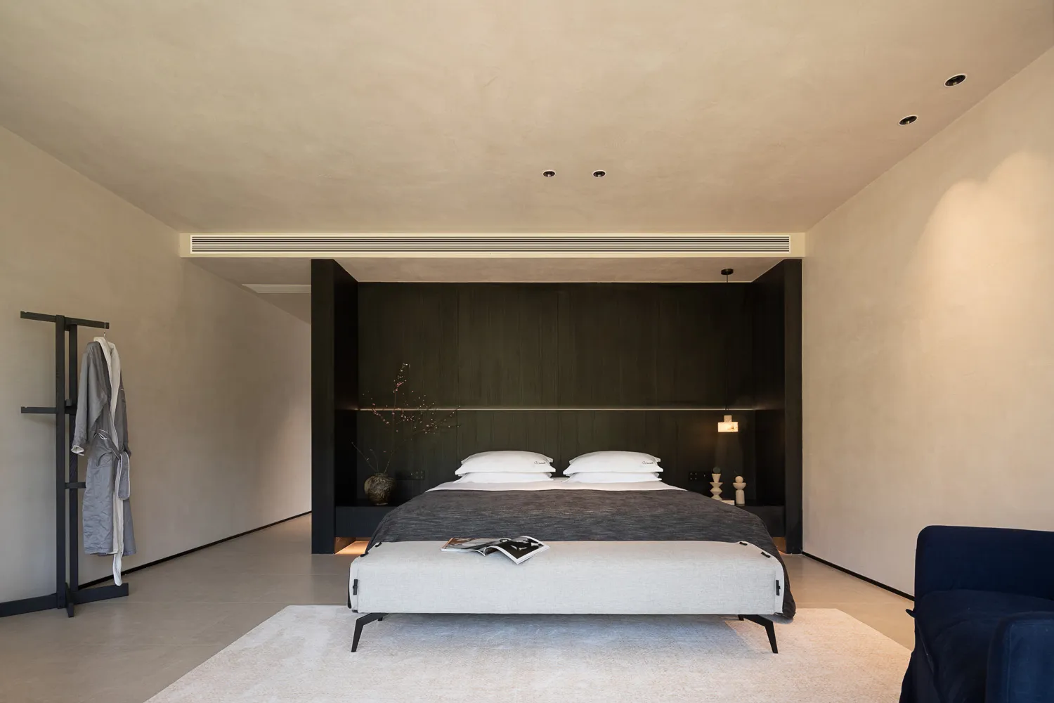 Modern minimalist bedroom with dark wood panel headboard wall, beige textured plaster walls, white bedding, upholstered bench, and blue velvet armchair