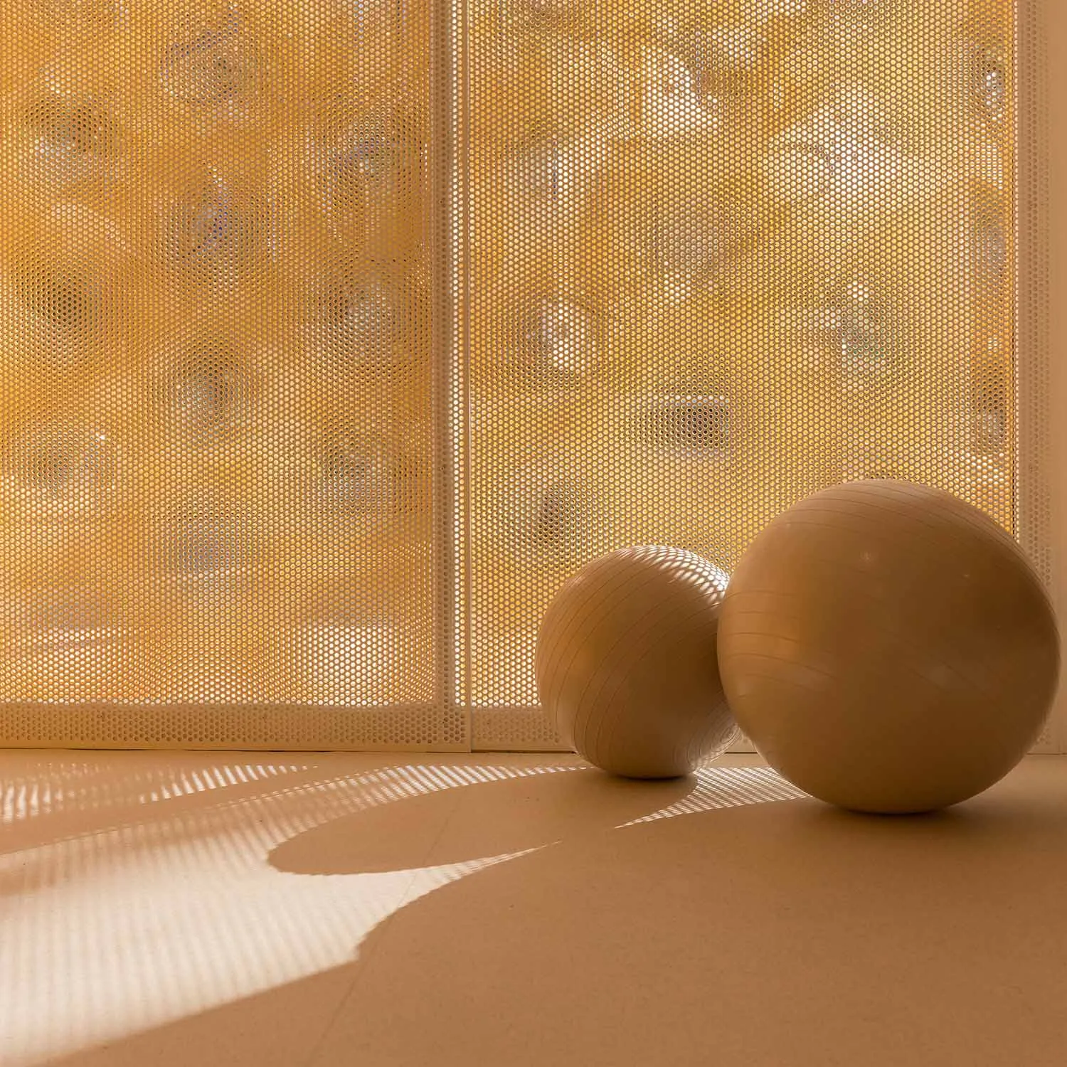 Two beige exercise balls rest on a light rubber floor in front of a glowing golden perforated screen that casts striped shadows.