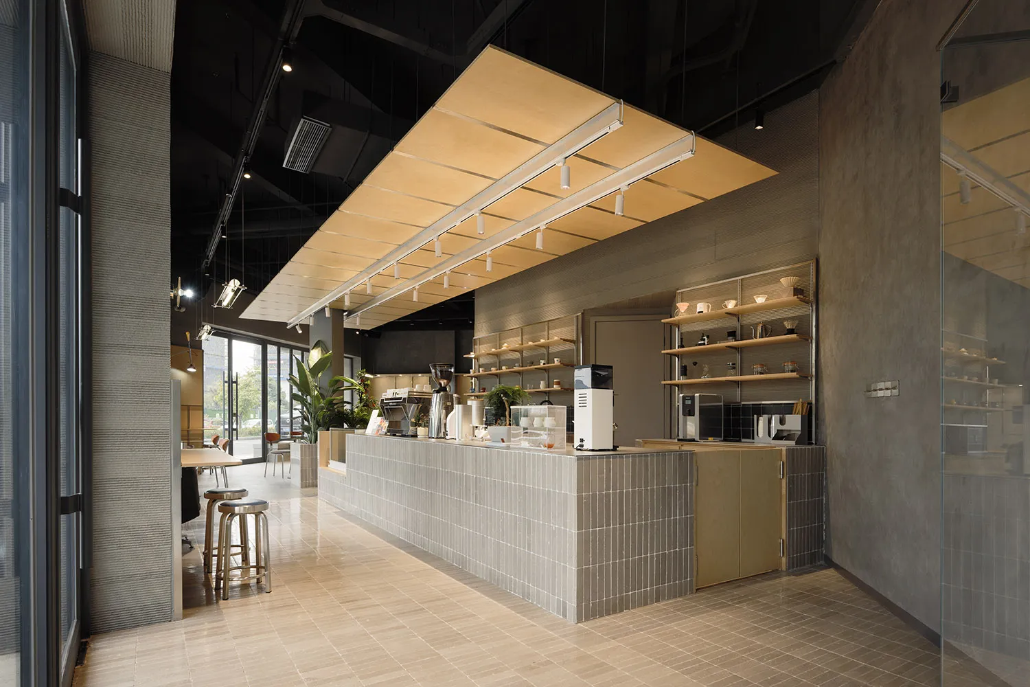 Modern minimalist cafe interior featuring a long grey concrete tile service counter, wooden canopy lighting fixture, metal bar stools, and open shelving against concrete walls.