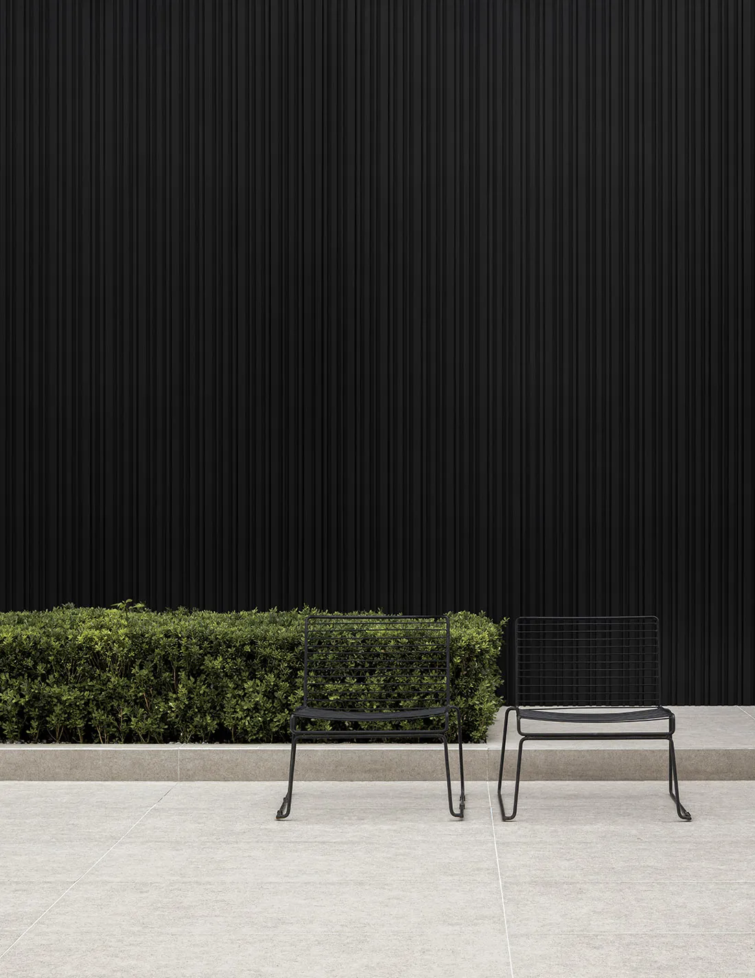Two black metal wire chairs sitting on a light grey tiled patio in front of a dark corrugated wall and a row of green bushes.