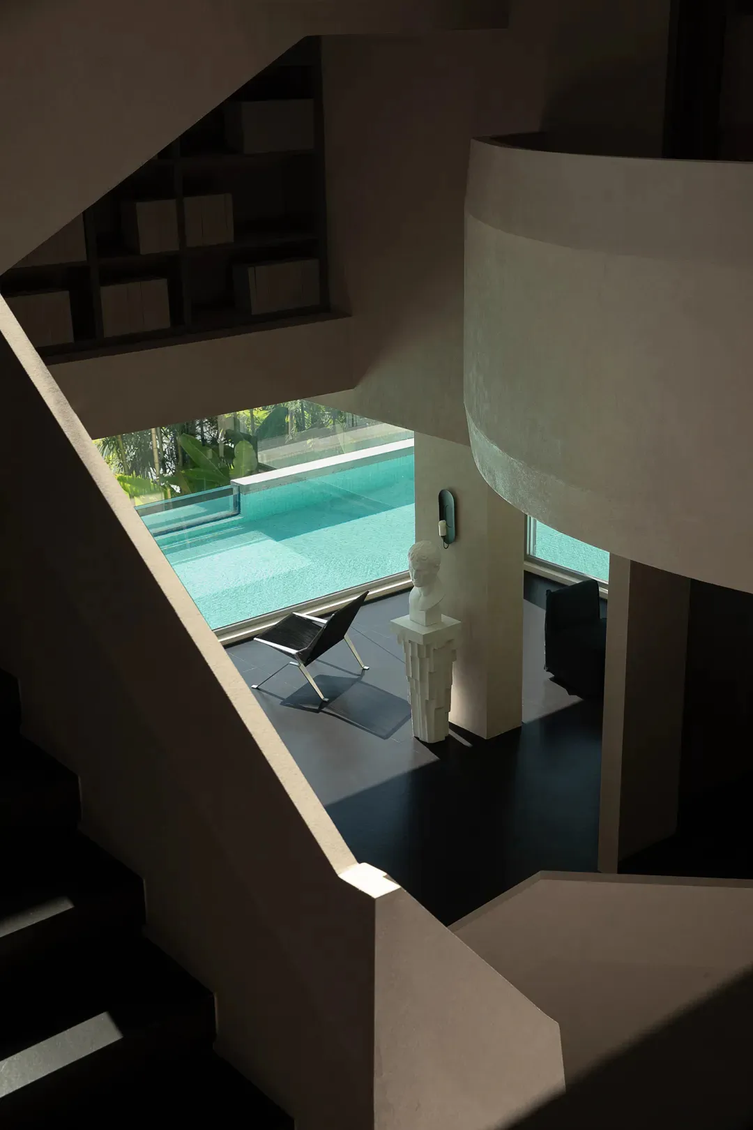 View from above of a modern curved concrete staircase overlooking a contemporary living area with a white classical bust on a pedestal, black leather chair, and large glass windows revealing an infinity pool surrounded by tropical greenery.