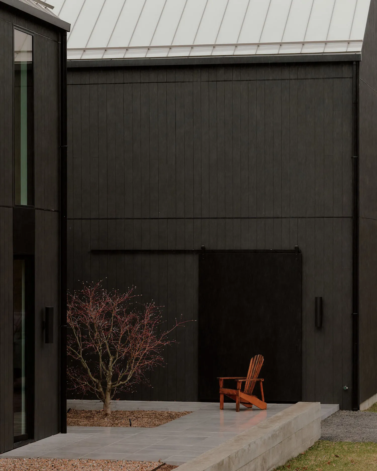 Modern black charred wood house exterior with concrete patio, small red-leaf tree, and orange Adirondack chair in a minimalist courtyard.