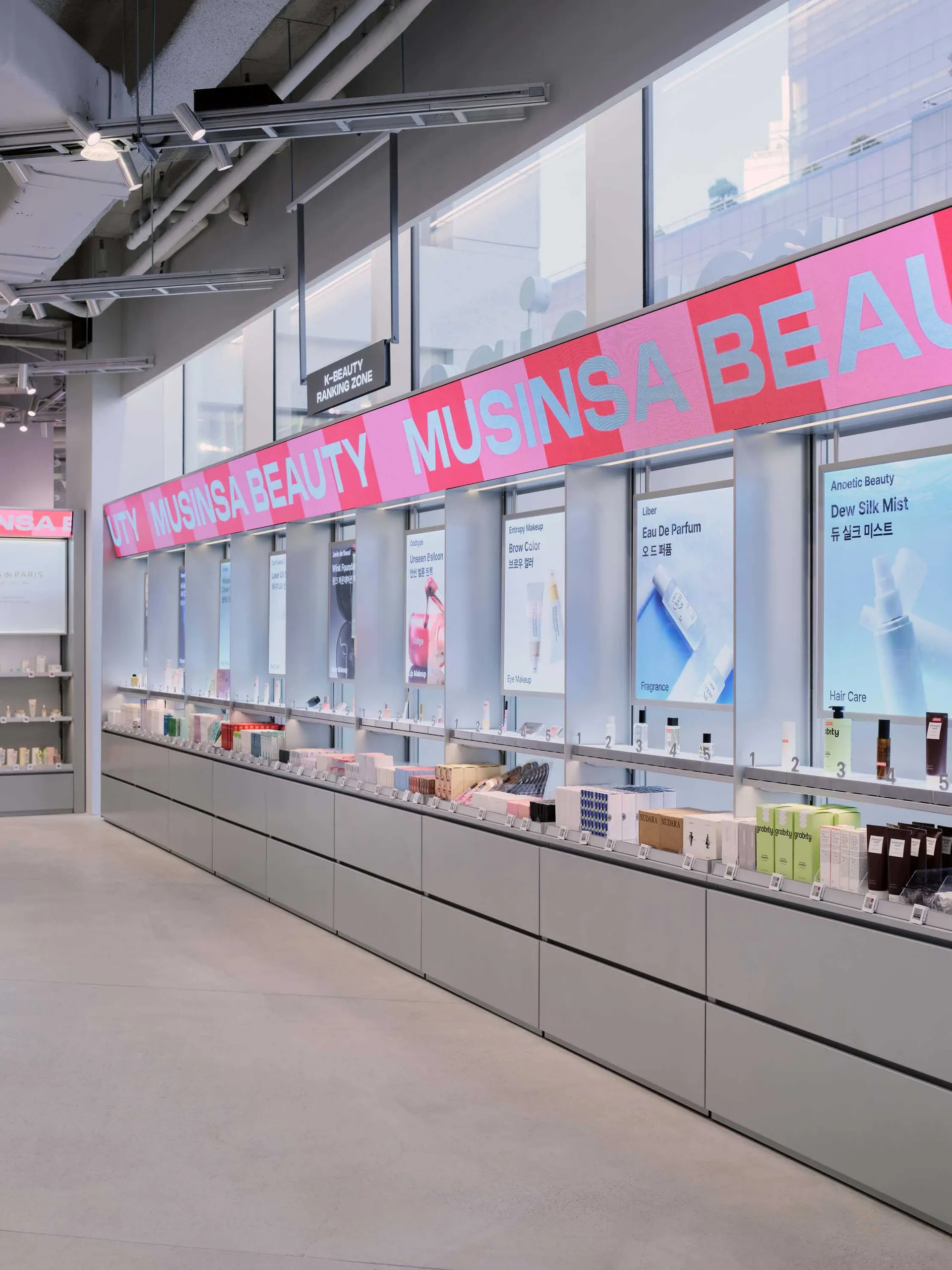 現代美妝店內的長型極簡零售展示牆,配有灰色櫃體、背光商品凹槽,以及大型粉紅色 MUSINSA BEAUTY 招牌。