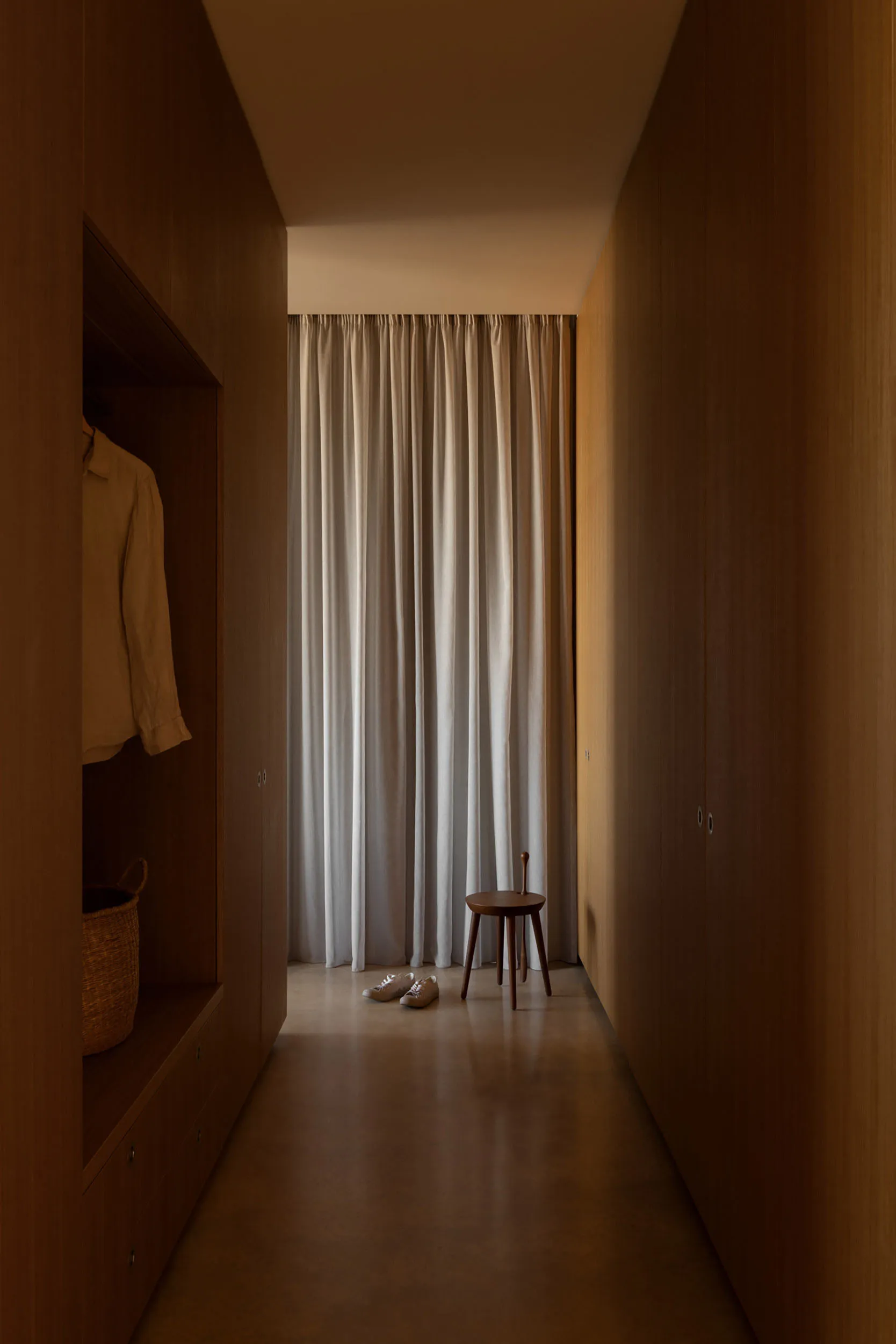 Narrow walk-in closet featuring floor-to-ceiling walnut wood cabinetry, a small wooden stool, white linen curtain, and woven basket storage.