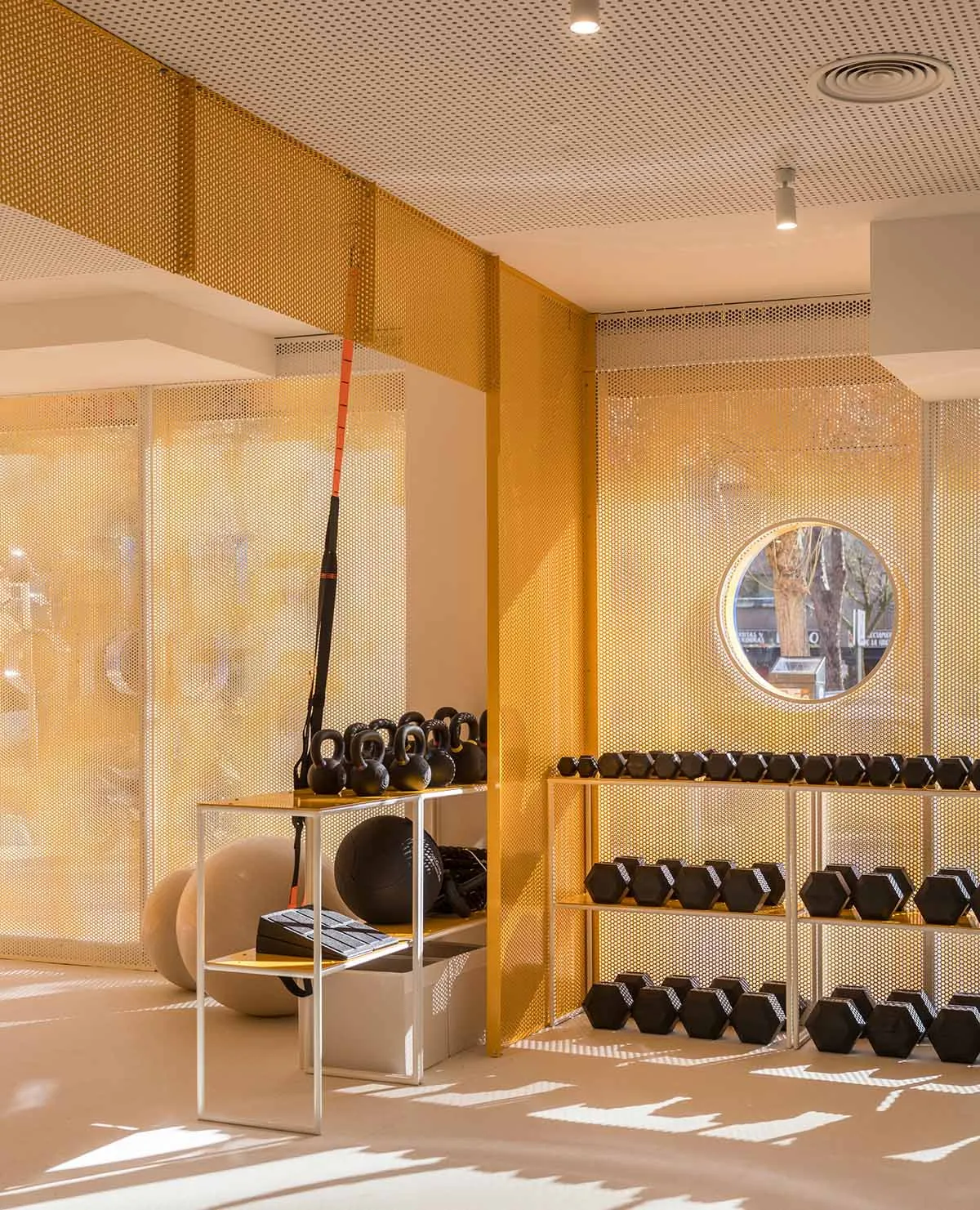 Modern home gym interior with golden perforated metal room dividers, dumbbell racks, kettlebells, exercise balls, and a circular window letting in natural light.