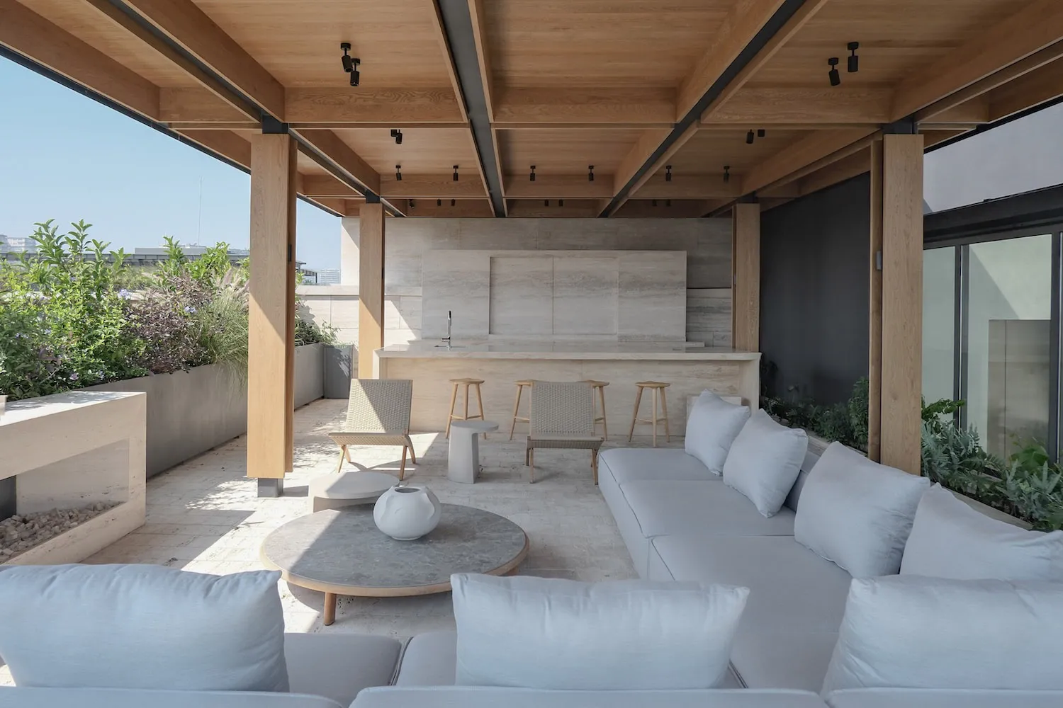 Modern rooftop terrace with light grey outdoor sofa, round travertine coffee table, built-in outdoor kitchen in travertine, wooden ceiling beams and abundant greenery under a clear blue sky.