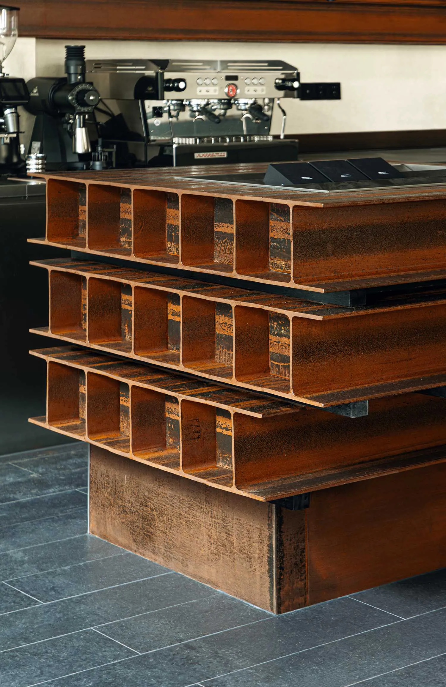 A sculptural coffee table made from stacked rusted H-beam steel sections sits on a dark tiled floor in front of a professional espresso machine.