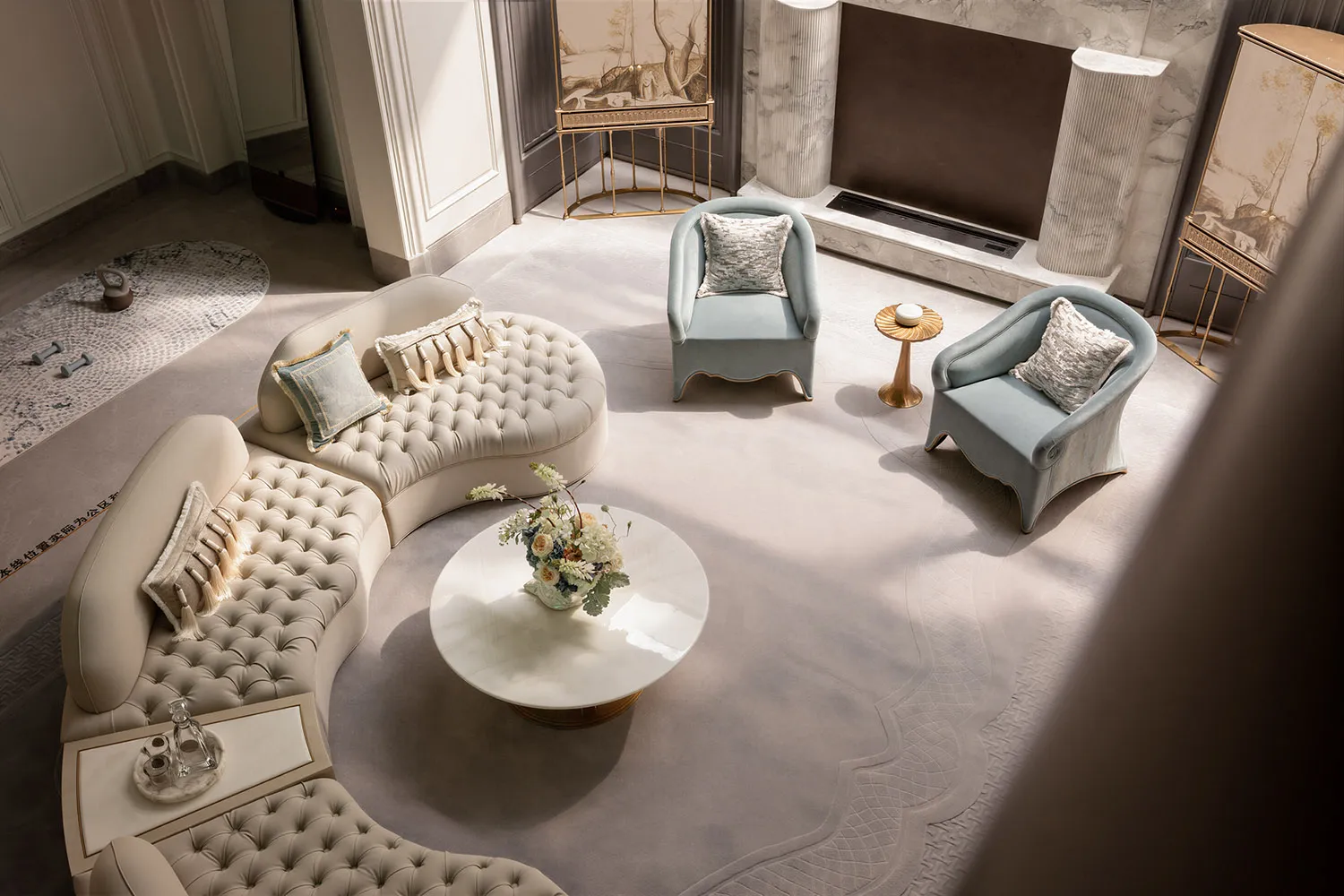 High-angle view of a luxurious living room with a large curved tufted cream sofa, two light-blue armchairs, a round white marble coffee table with floral arrangement, and marble fireplace surround.