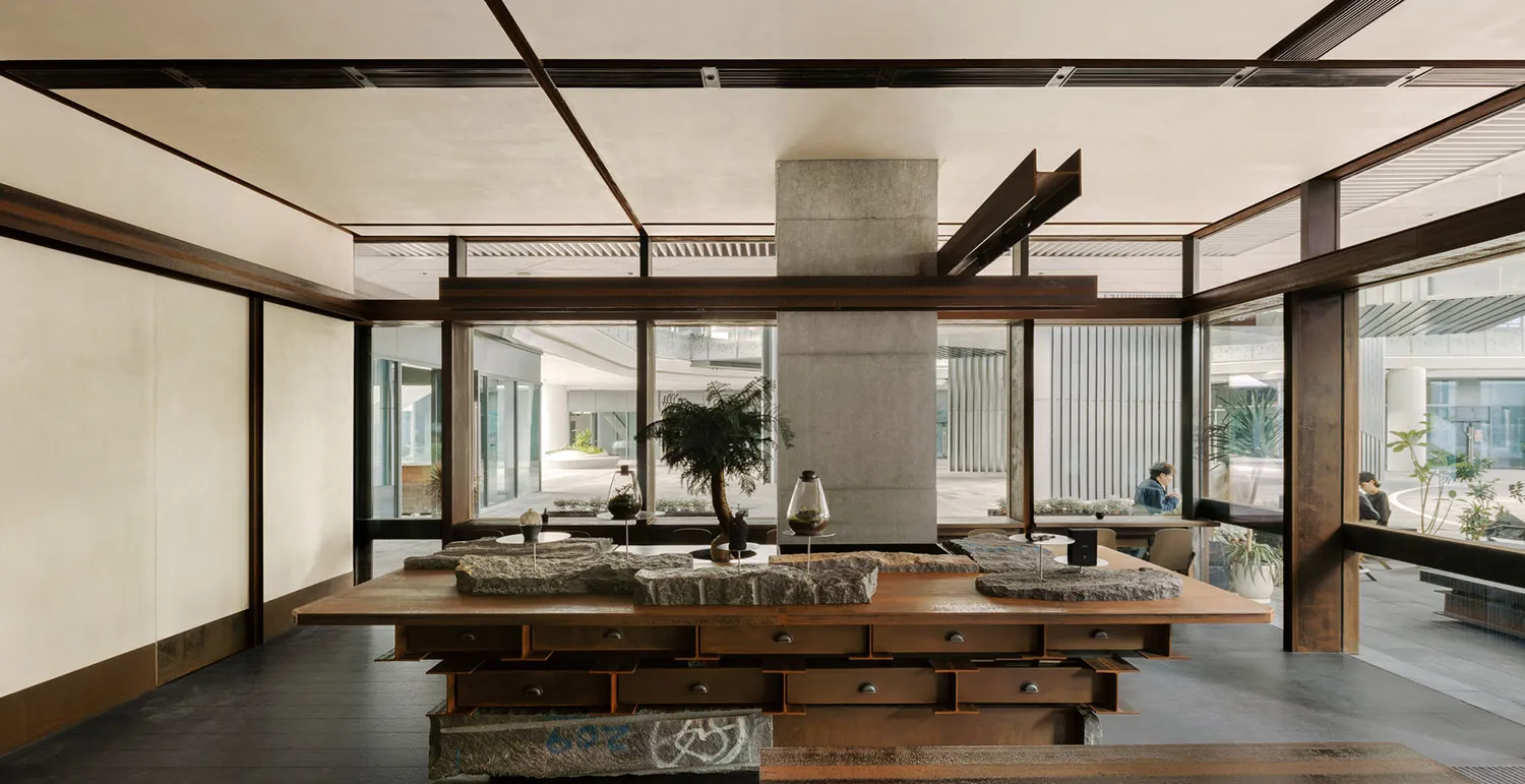 Spacious modern design studio interior featuring a large reclaimed wood worktable with stone models, a central concrete structural pillar, dark metal beams, and large glass windows overlooking a garden courtyard.