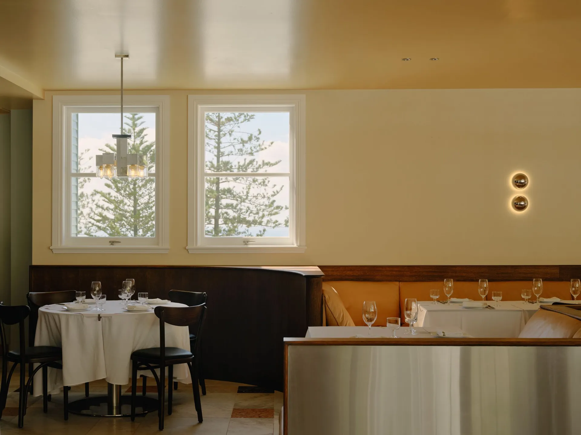 Elegant restaurant interior featuring dark walnut wainscoting, a bright orange upholstered banquette, round tables with white linens, dark wood chairs, and two large windows overlooking pine trees and the ocean.