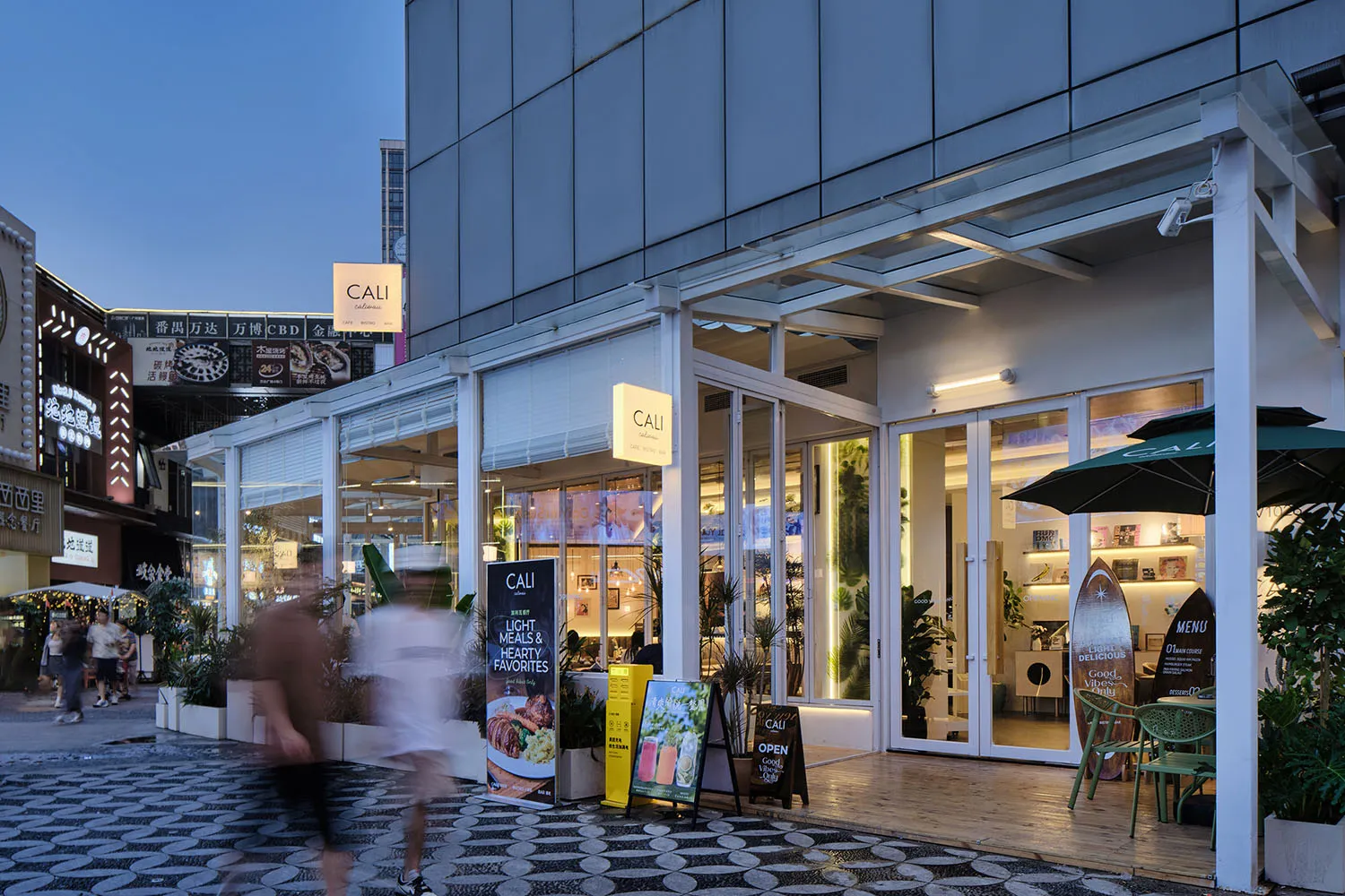Modern cafe named CALI with large glass windows, white structural frames, outdoor seating, and potted plants along a patterned stone pedestrian street at dusk.