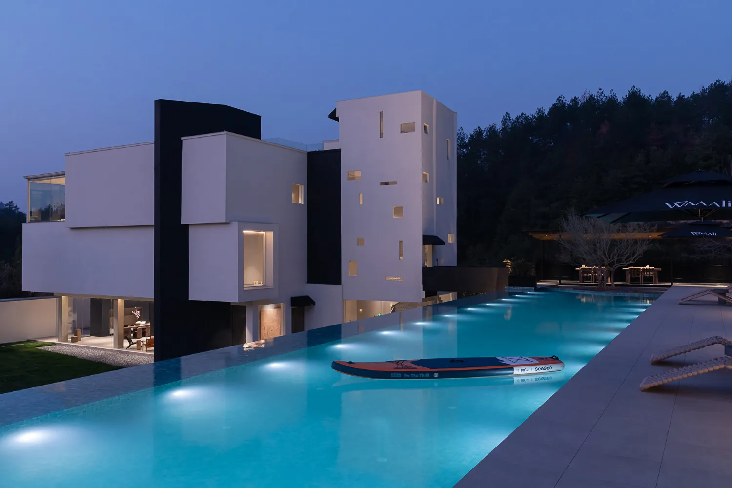 Modern white concrete villa with black architectural accents and a long illuminated infinity pool at dusk, with lounge chairs and forested hillside in the background.
