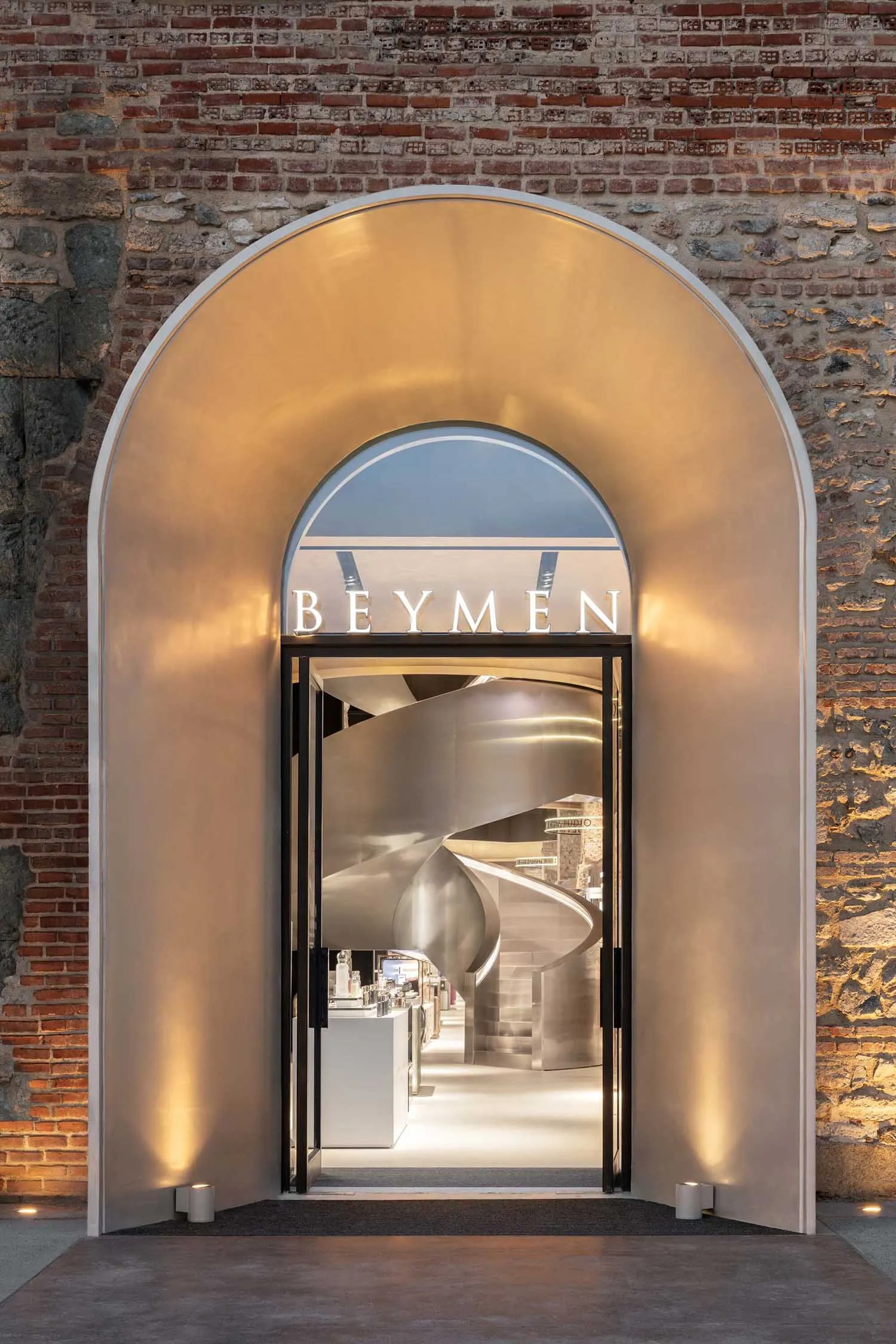 View of a modern luxury store entrance featuring a large illuminated stainless steel arch set within an old brick wall, revealing a dramatic spiral staircase inside.