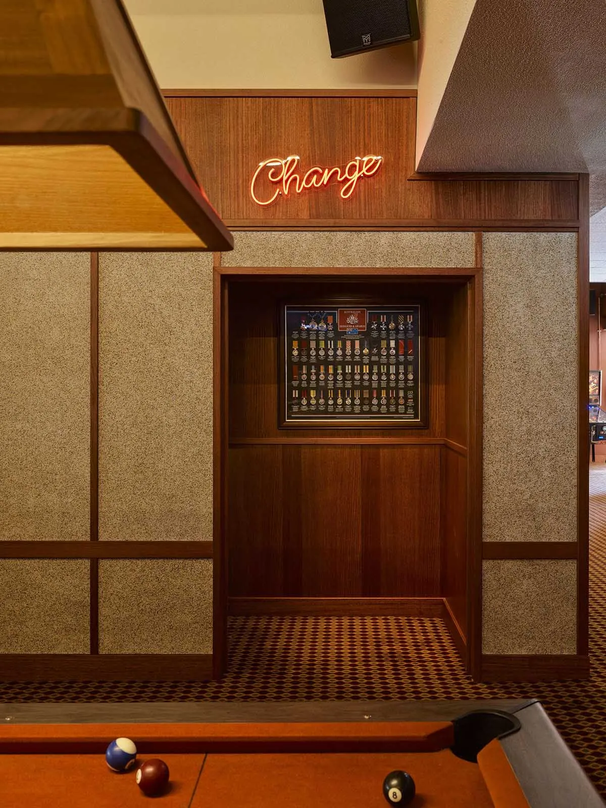 Interior view of a classic billiard room with walnut wood paneling, textured walls, a neon sign, and a framed display of military medals above a patterned carpet.