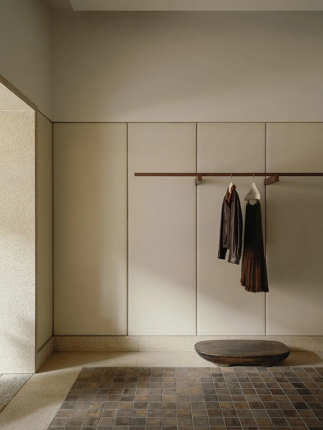 Minimalist entryway with beige terrazzo wall, built-in cabinetry, wooden clothing rack holding a leather jacket and brown dress, and a dark live-edge wooden bench on a brown tiled floor.
