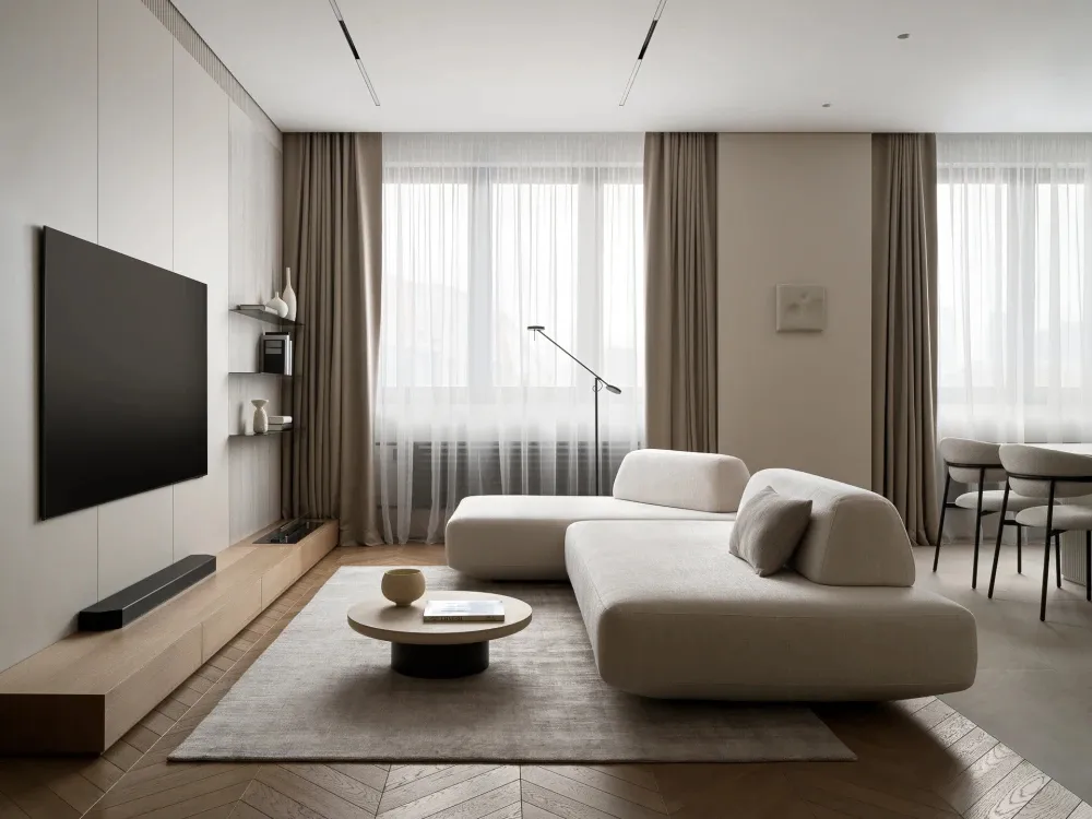 Modern minimalist living room featuring a large L-shaped cream modular sofa, round oak coffee table on a textured rug, wall-mounted TV above a light oak entertainment console, and floor-to-ceiling sheer curtains allowing natural light to flood the space.