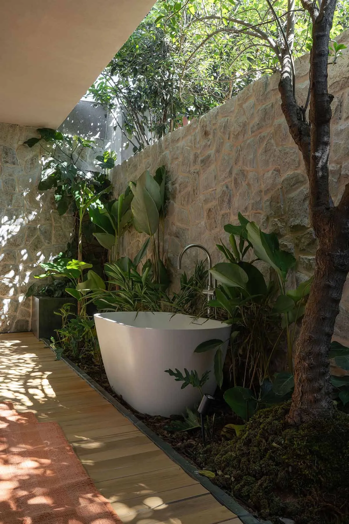 Modern freestanding white bathtub placed in an outdoor garden surrounded by lush green plants, stone walls, and a wooden deck under natural sunlight.