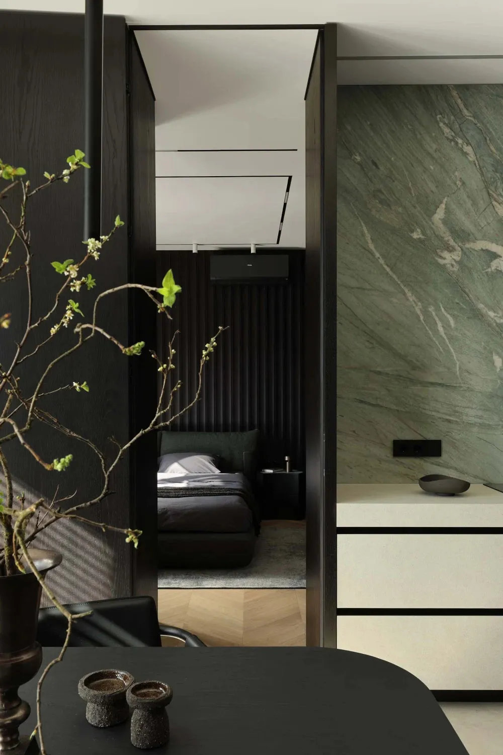 View through a dark wood doorway into a modern bedroom with a large green marble accent wall, dark upholstered bed, and herringbone wood flooring.