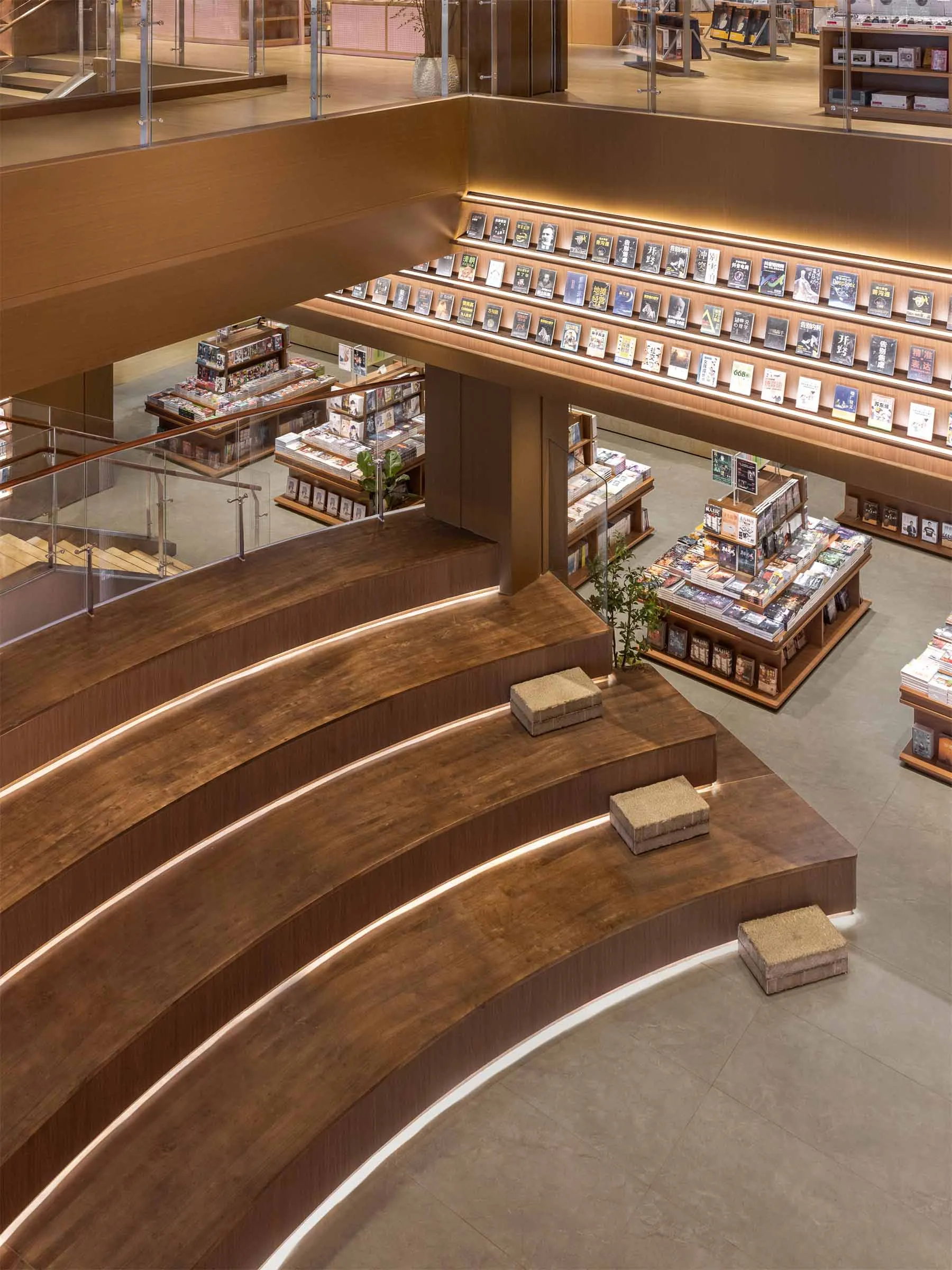 View from above of a curved wooden staircase with integrated LED lighting leading to the second floor of a spacious, modern bookstore with extensive wooden bookshelves and display tables.