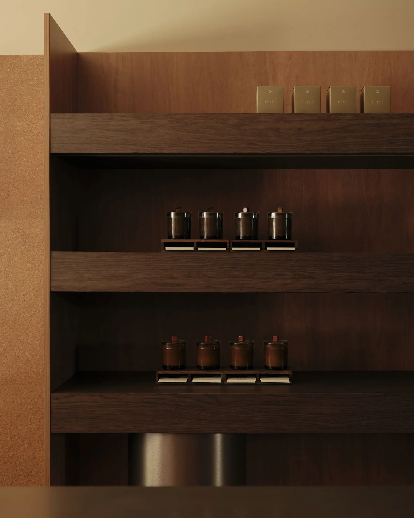 Close-up of a minimalist built-in walnut wood shelving unit featuring neatly arranged dark glass tea canisters with wooden lids on two open shelves, with matching product boxes displayed on the top shelf against a warm cork wall.