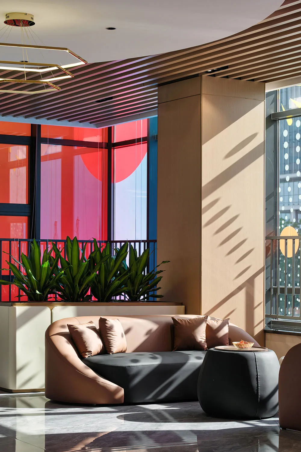 Modern lobby lounge featuring a curved two-tone leather sofa, dark ottoman, wooden slat ceiling, and large windows with red circular art installation.