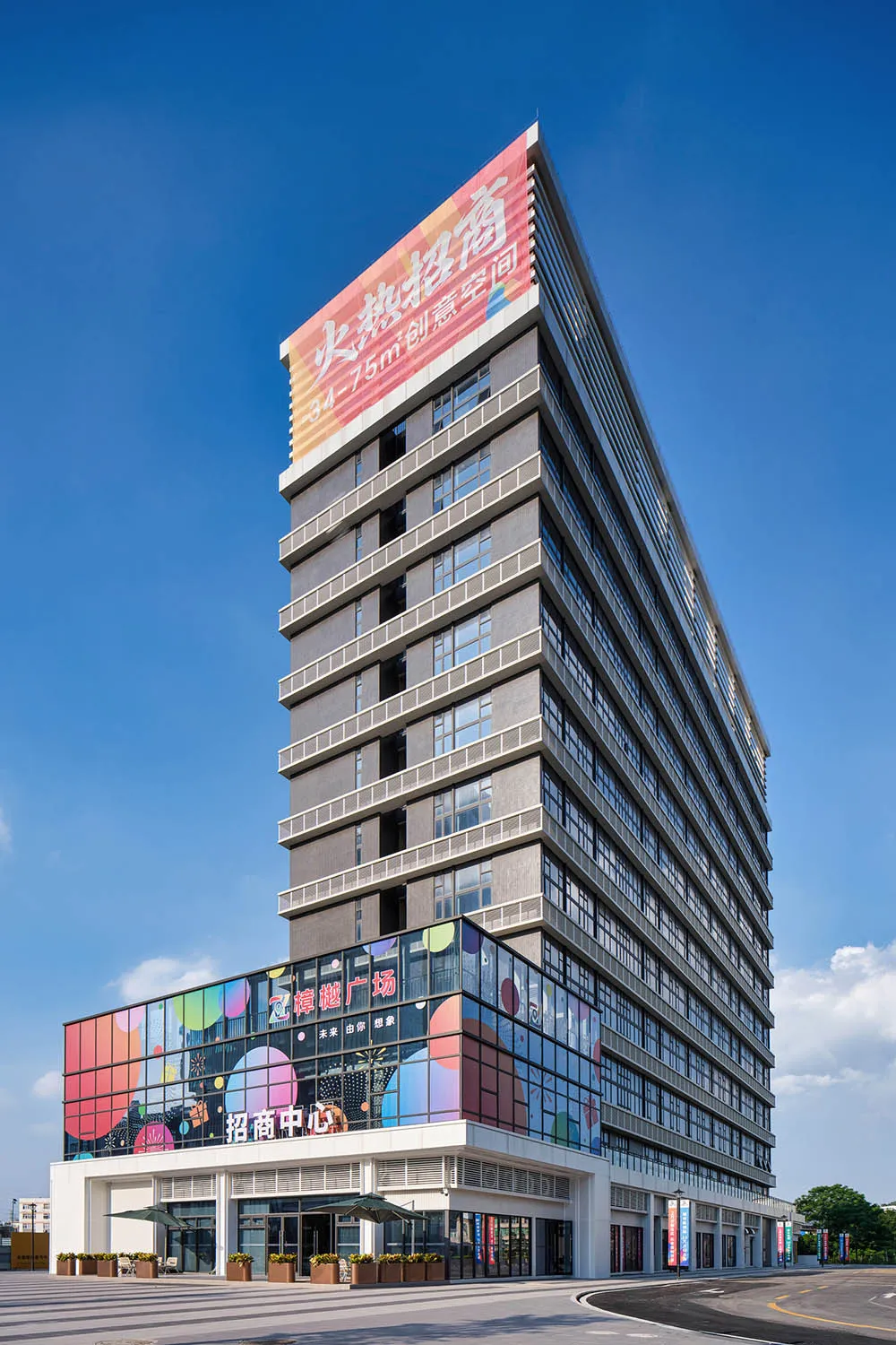 Exterior view of a tall modern office building with a colorful glass facade on the lower levels and large Chinese signage, set against a clear blue sky.