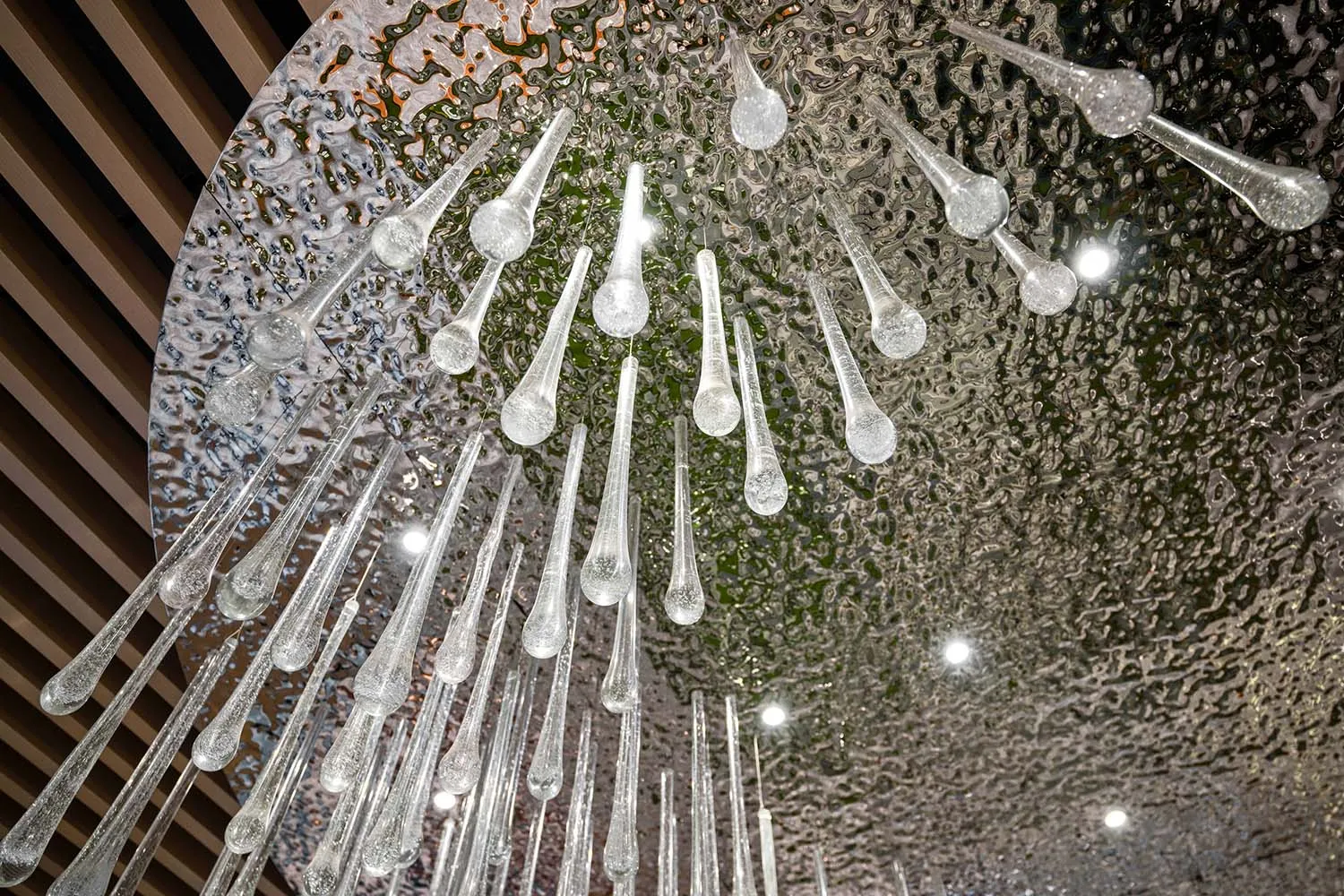 Close-up view looking up at an artistic chandelier featuring numerous clear glass teardrop and rod elements hanging from a large circular hammered silver ceiling panel with integrated spotlights.