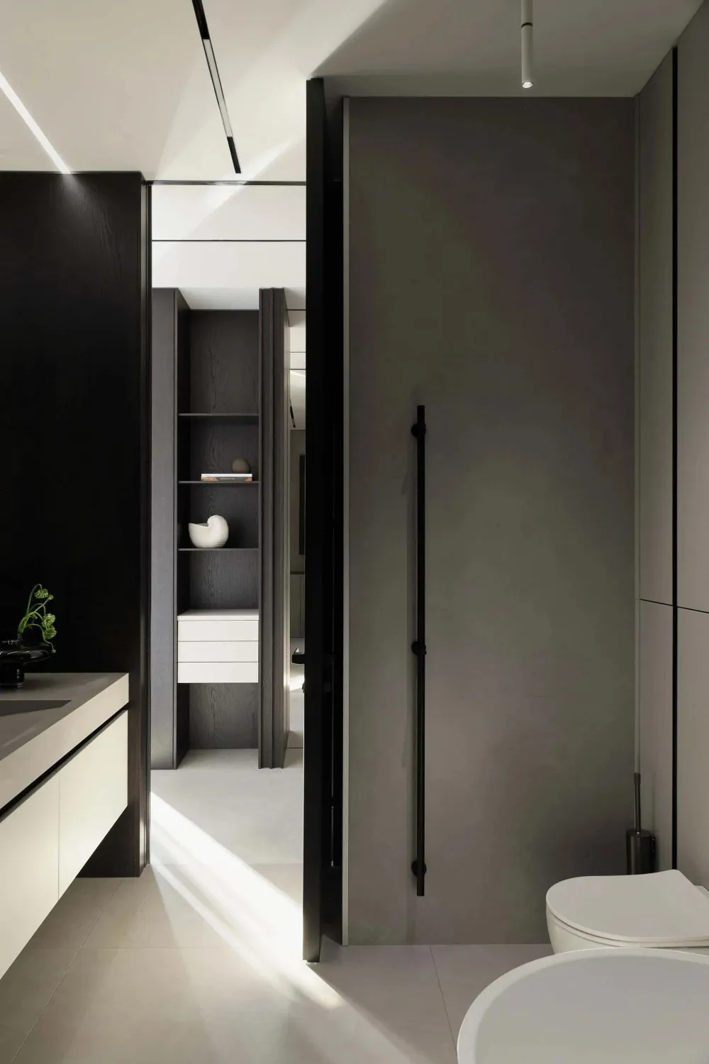 Modern minimalist bathroom interior with dark wood cabinetry, white floating vanity, built-in open shelving, matte black door, and white ceramic toilet under dramatic natural light.