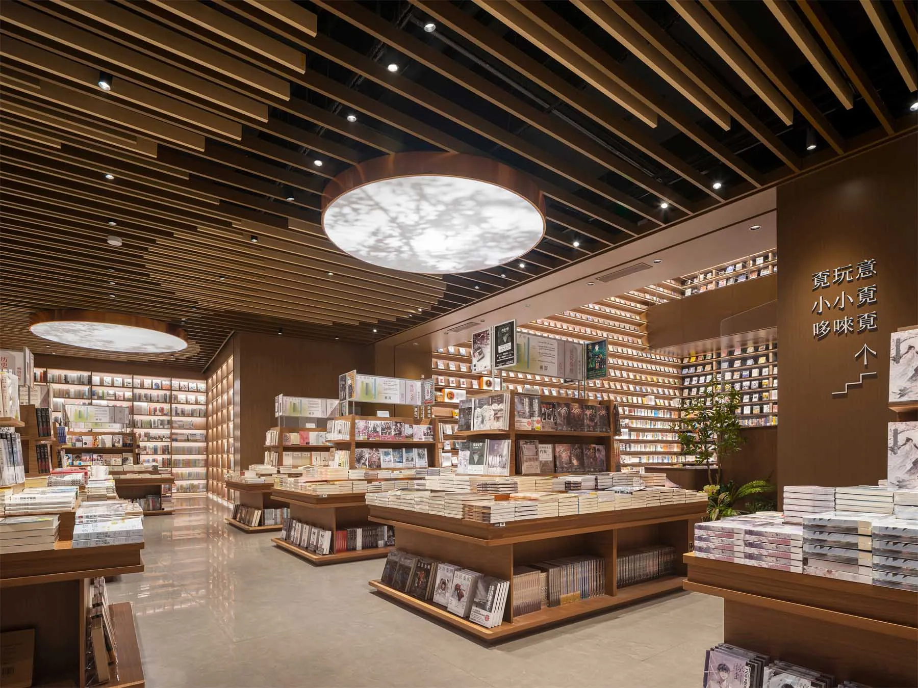 Modern bookstore interior with extensive wooden slat ceiling, large circular pendant lights casting leaf patterns, and bookshelves filled with books.