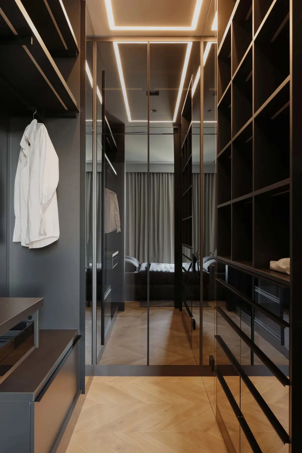 Modern dark walk-in closet with mirrored wardrobe doors, herringbone oak flooring, and integrated LED ceiling lighting, white shirt hanging on the left.